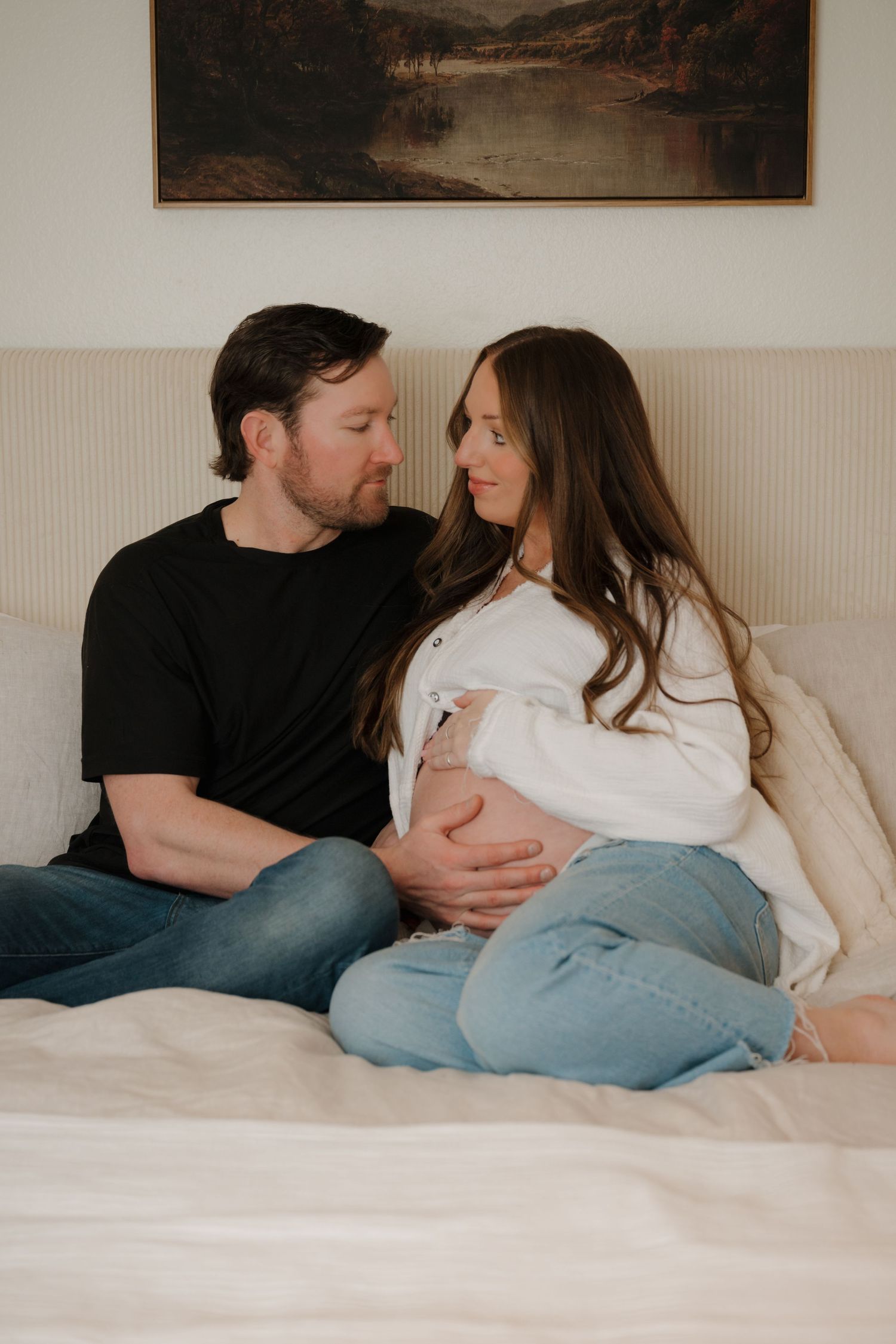 Cozy indoor maternity photo with couple relaxing on white couch beneath landscape artwork.