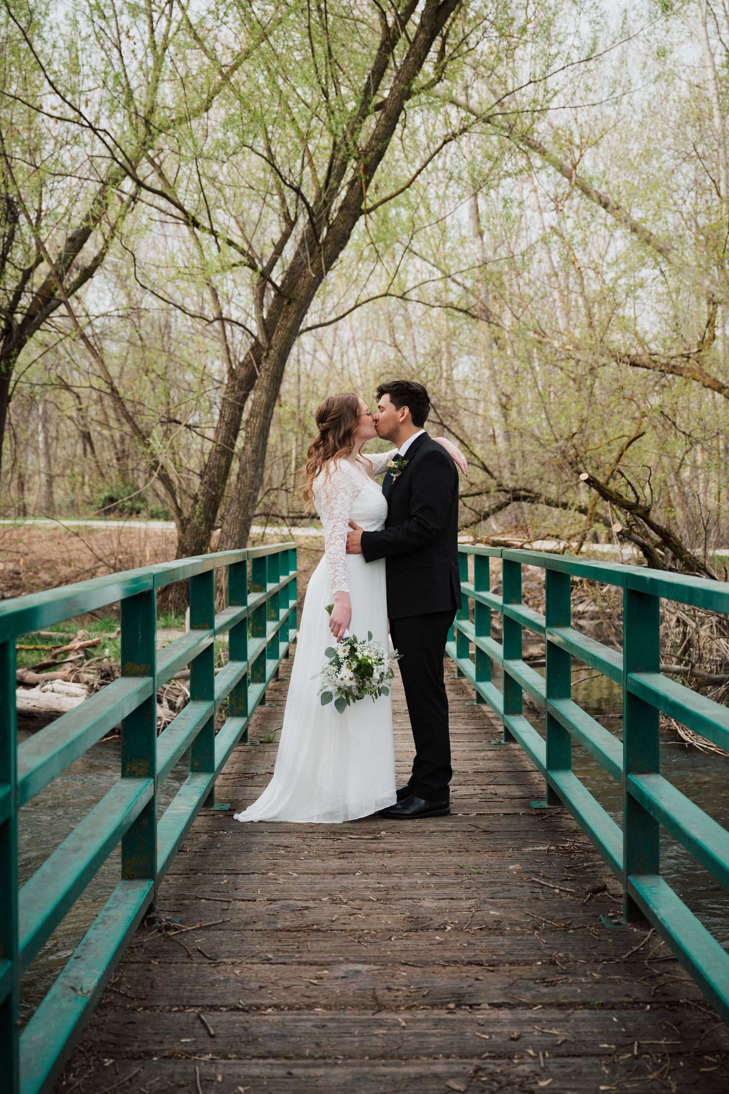 A couple in wedding attire shares an intimate moment on a turquoise-railed wooden bridge surrounded by spring trees.