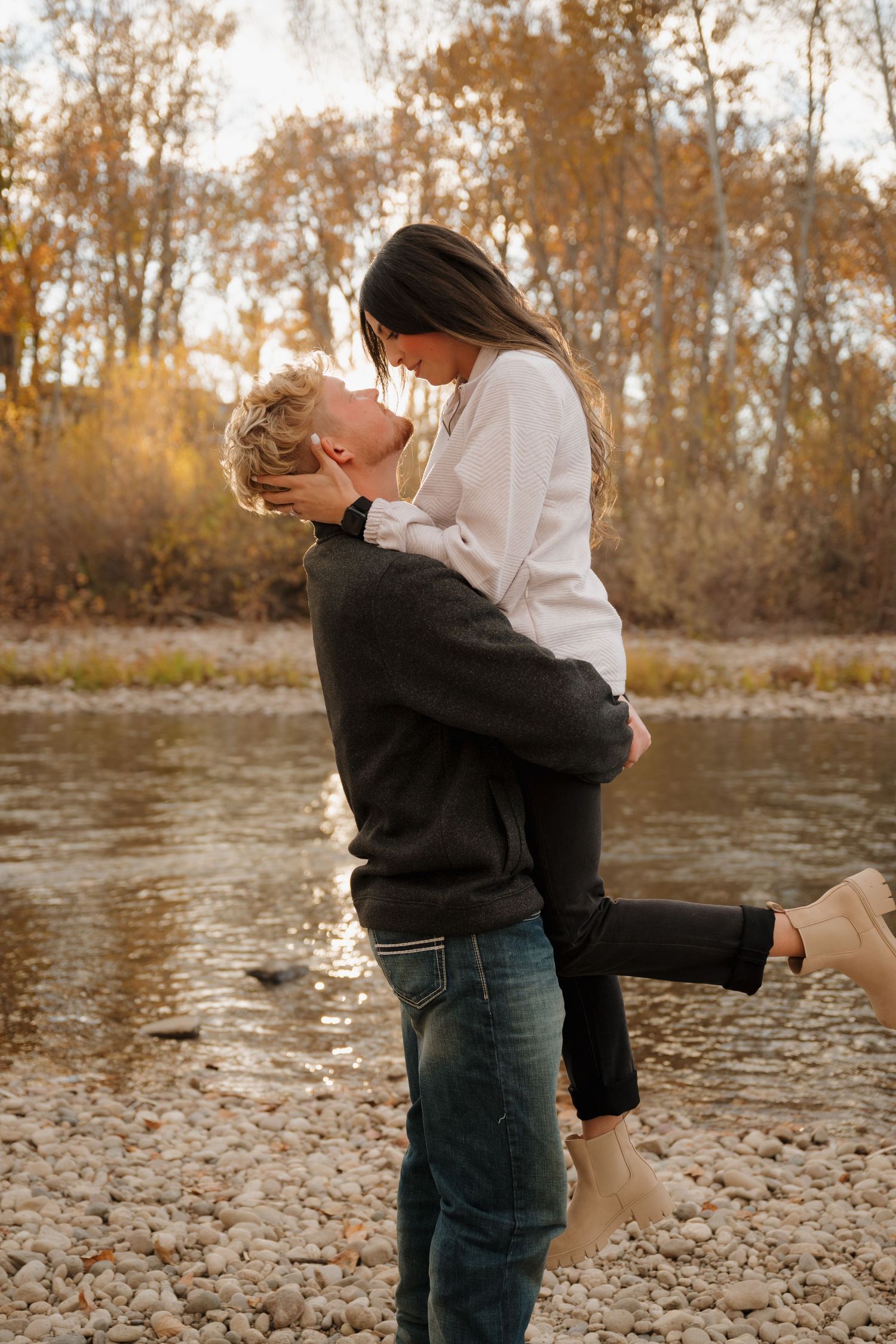 A romantic couple embraces by a riverbank during golden hour in autumn.