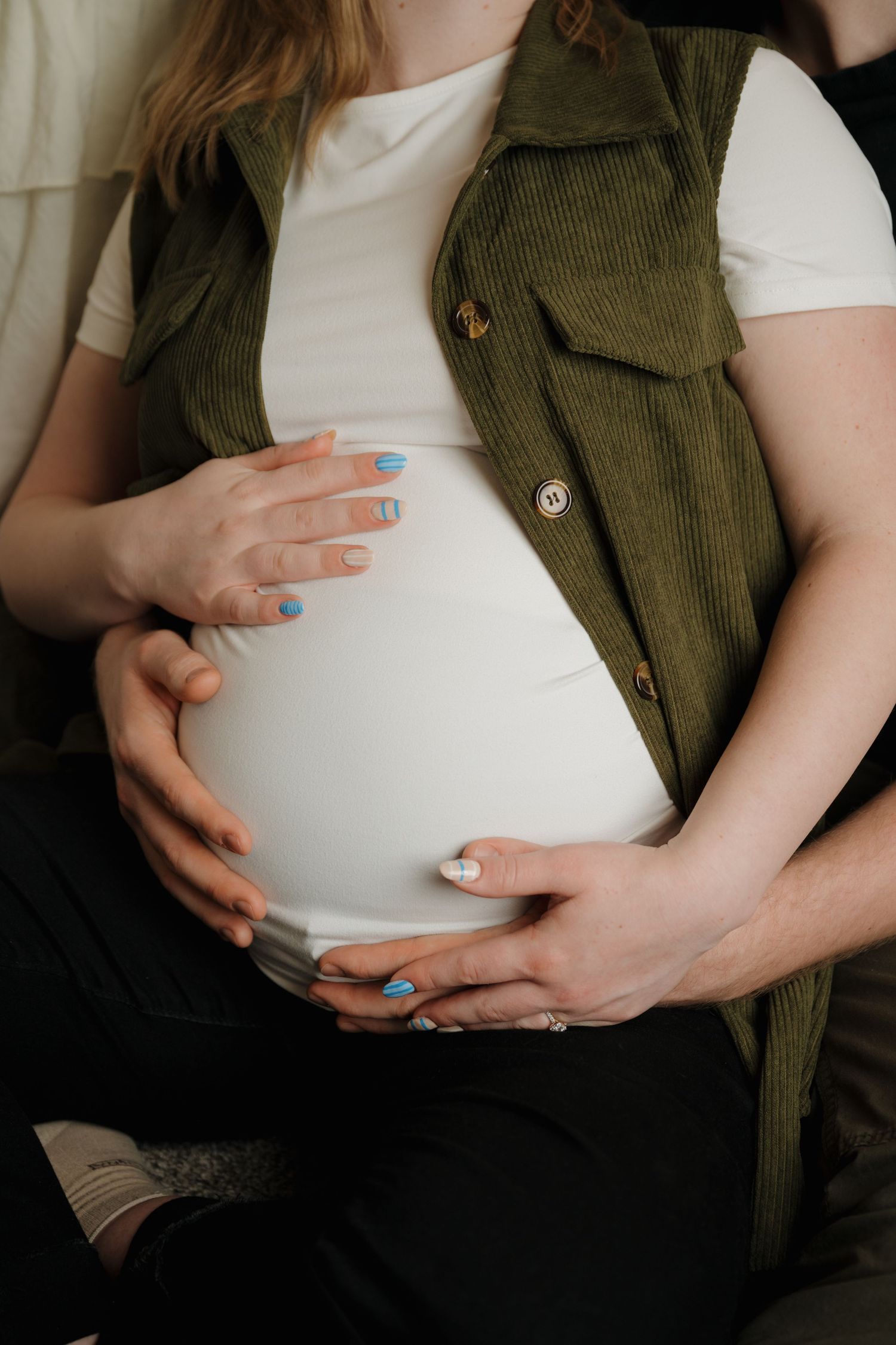 Hands gently resting on a pregnant belly, wearing a green vest and white shirt.