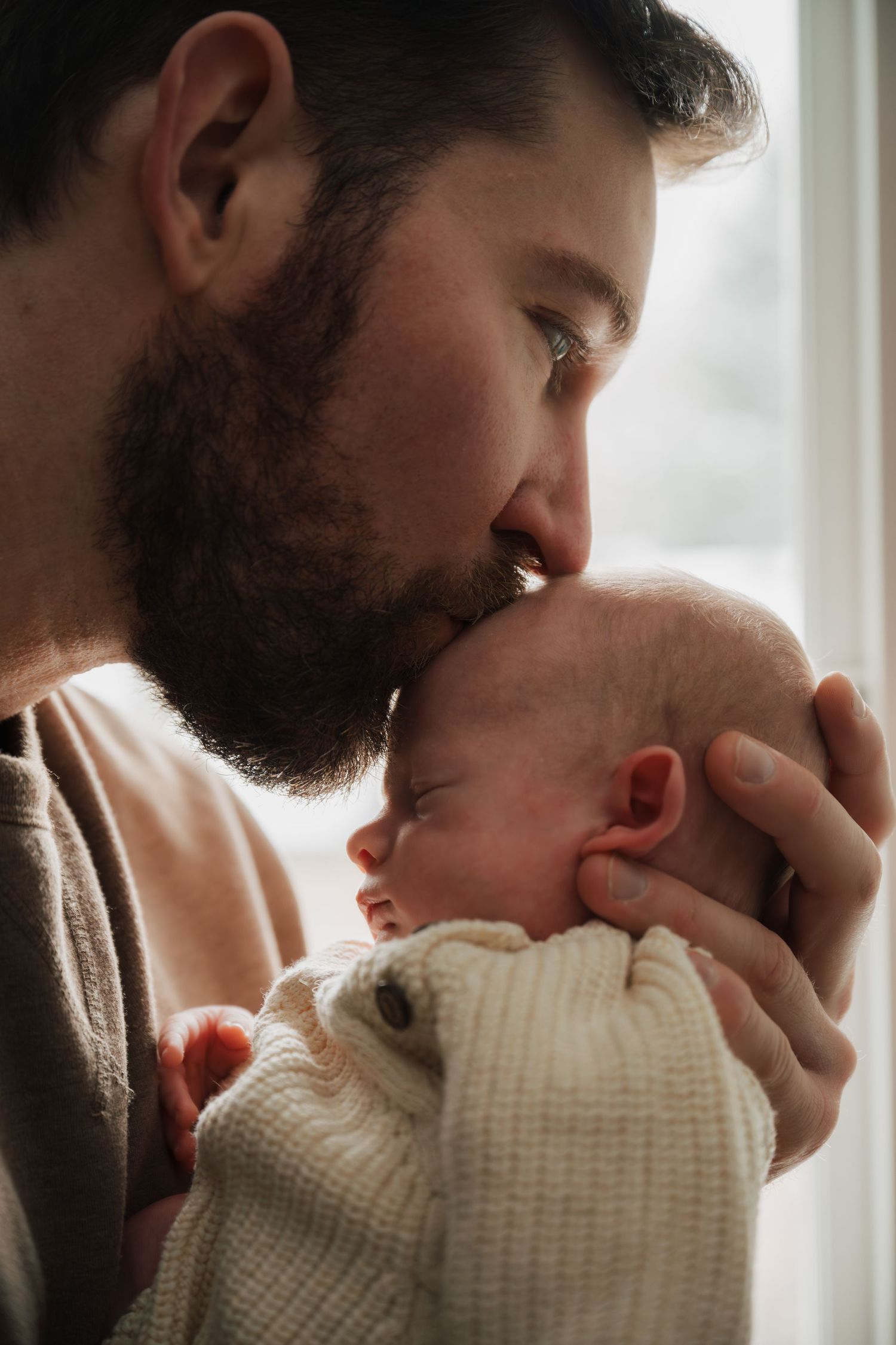 Parent in beige sweater tenderly kisses sleeping newborn wrapped in white knit blanket.
