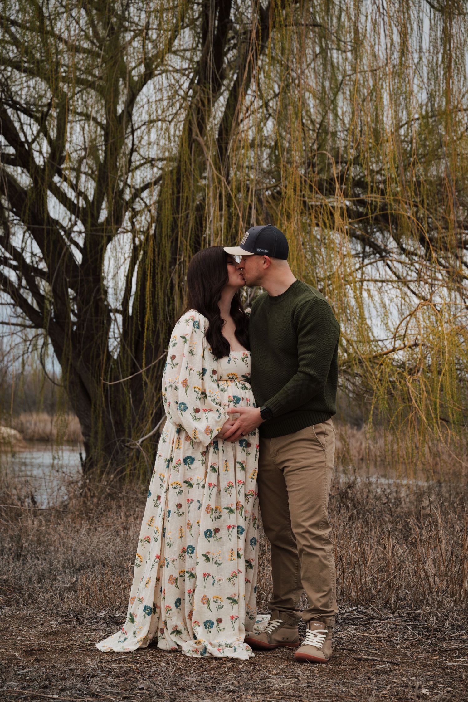 Romantic maternity photo of couple under weeping willow tree in winter landscape.