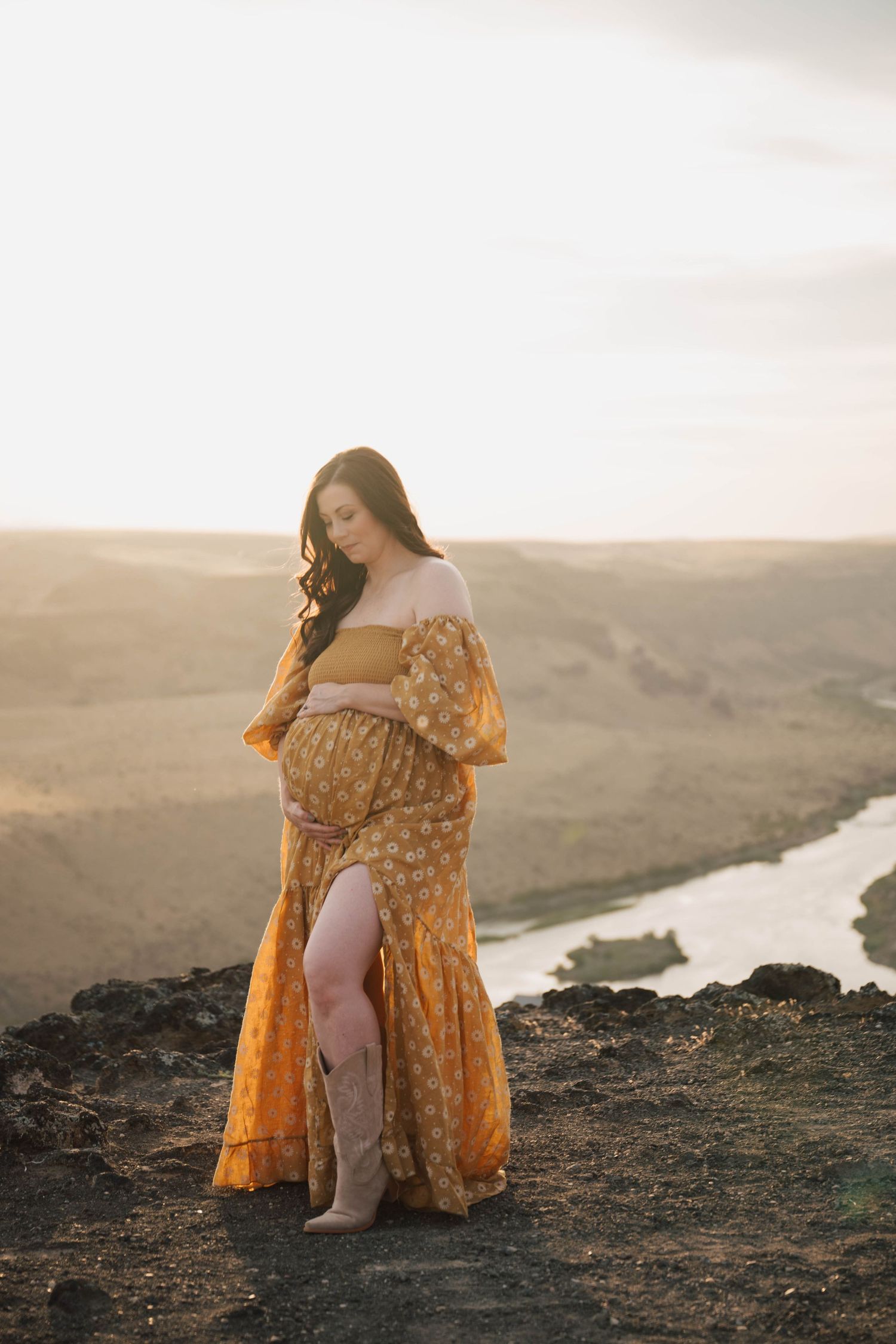 A person in a flowing yellow polka dot dress poses on rocky terrain at sunset with a river valley in the background.
