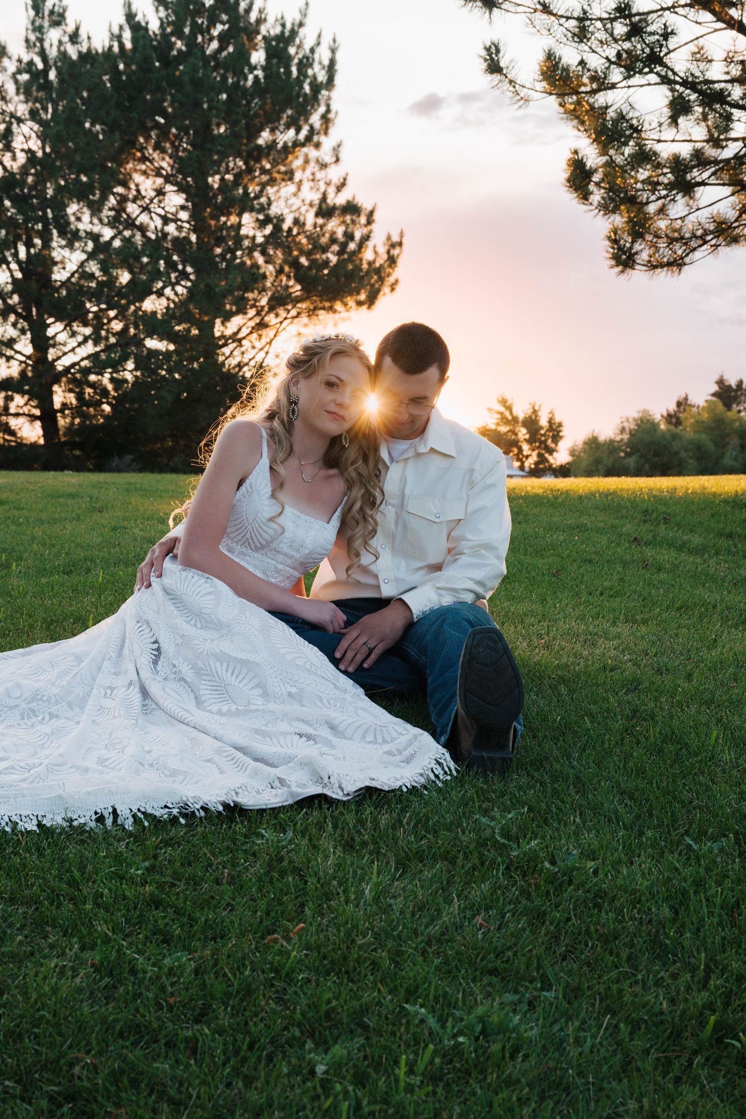 A couple sits together on grass during sunset as golden light filters through pine trees in the background.