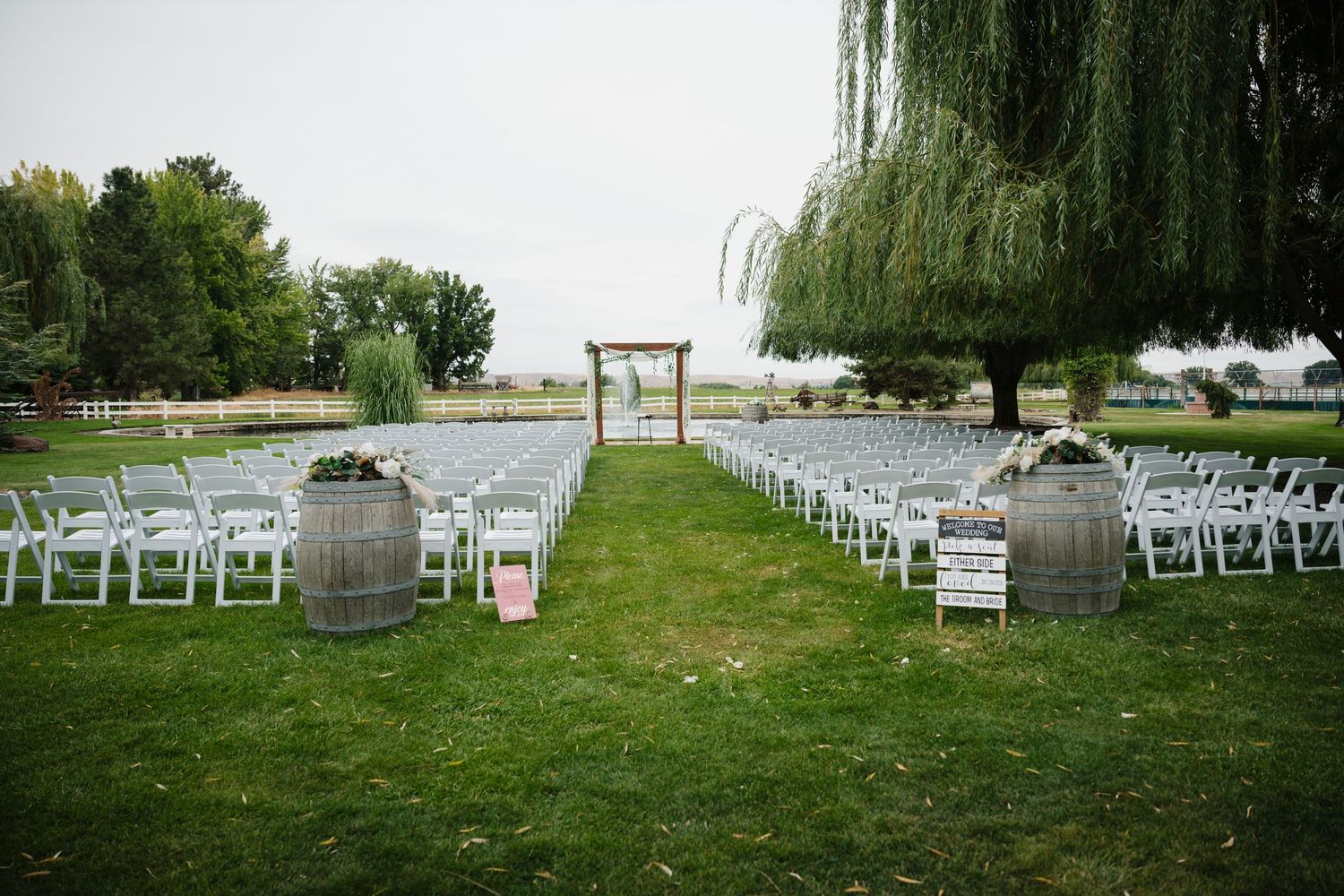 Outdoor wedding ceremony setup with white chairs and wooden arch beneath weeping willow trees by a lake.