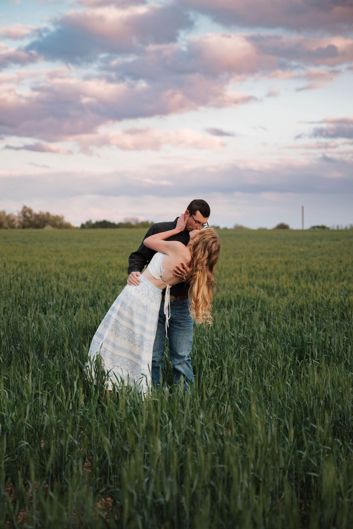 A romantic couple embraces in a lush green field during sunset with pink clouds overhead.