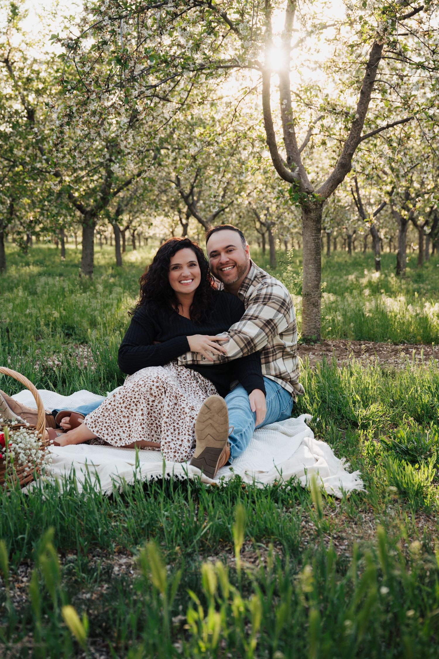 Couple enjoying a romantic spring picnic on white blanket in blooming orchard at sunset.