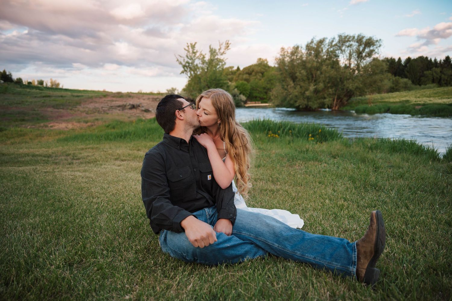 A couple sits together on grassy riverbank during sunset sharing an intimate moment in nature.
