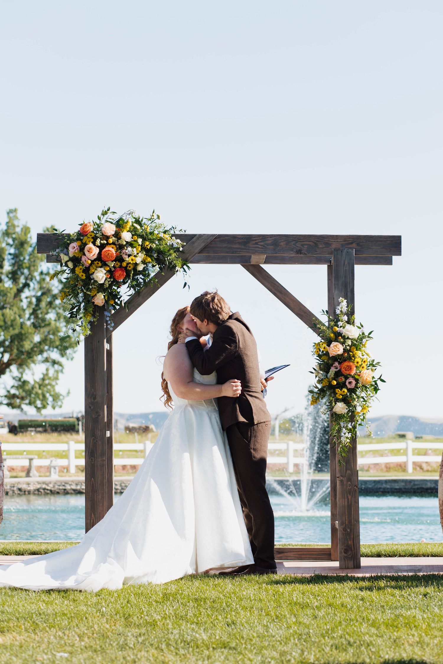 A romantic outdoor wedding ceremony takes place under a wooden pergola decorated with vibrant floral arrangements.