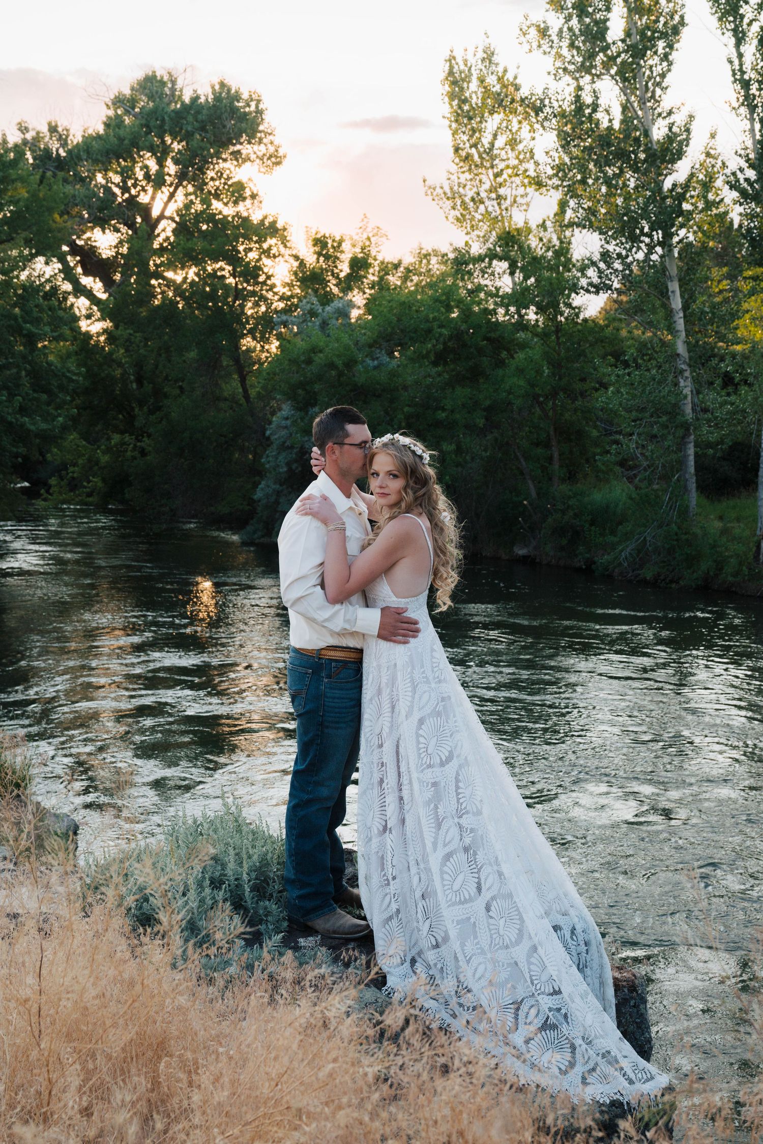 Two people share a romantic moment at sunset by a river, with a flowing white dress and rustic country setting.