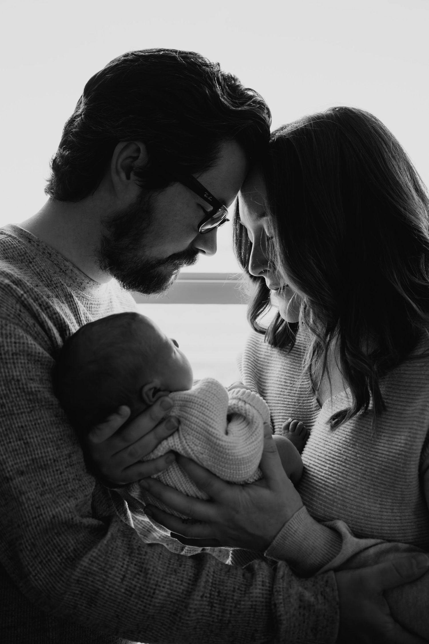 Intimate black and white photo of parents sharing tender moment while holding their newborn baby by a window.