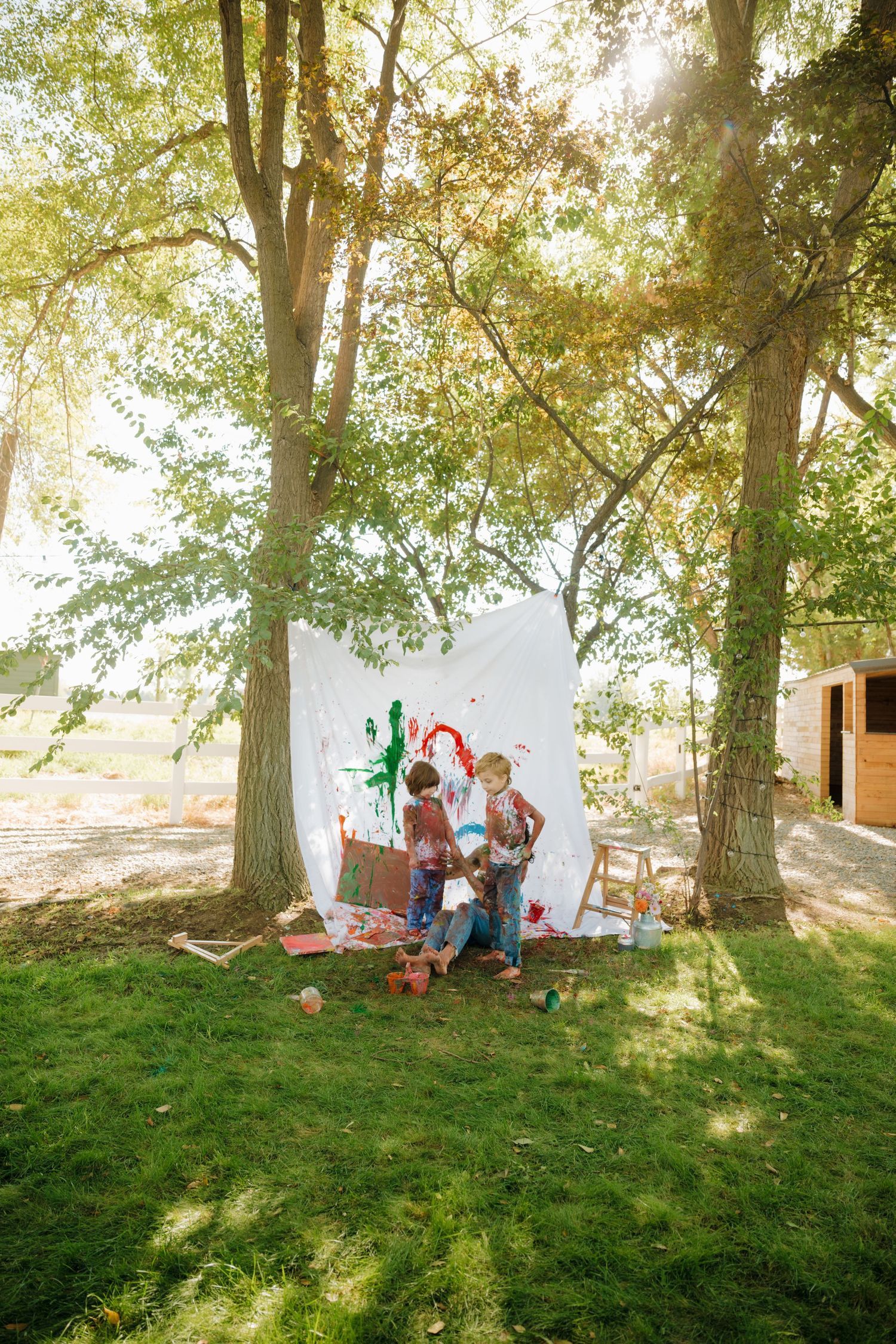 Family creating colorful paint splatter art on white canvas outdoors under tall trees.