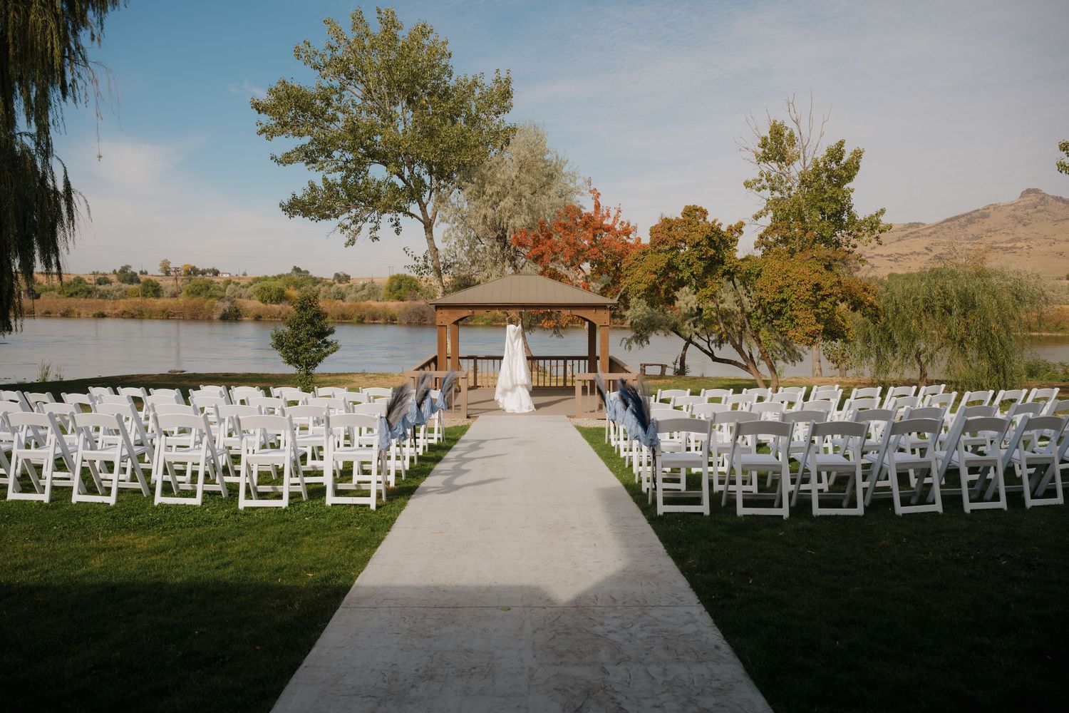 Outdoor wedding ceremony setup with white chairs and lakeside gazebo surrounded by autumn trees and mountains.