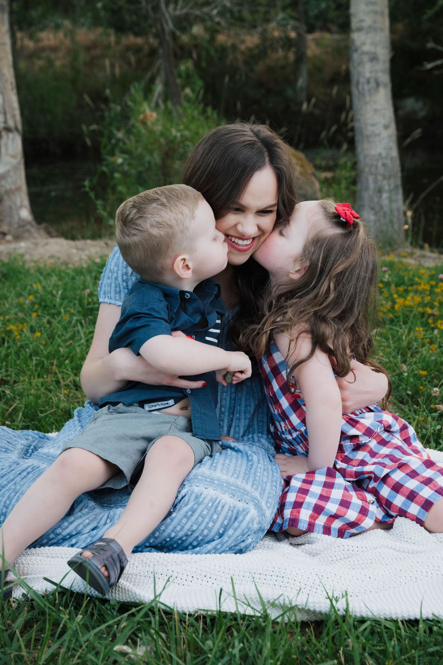 Sweet family moment captured on a blanket outdoors with two young children sharing kisses with their mother on a sunny day.
