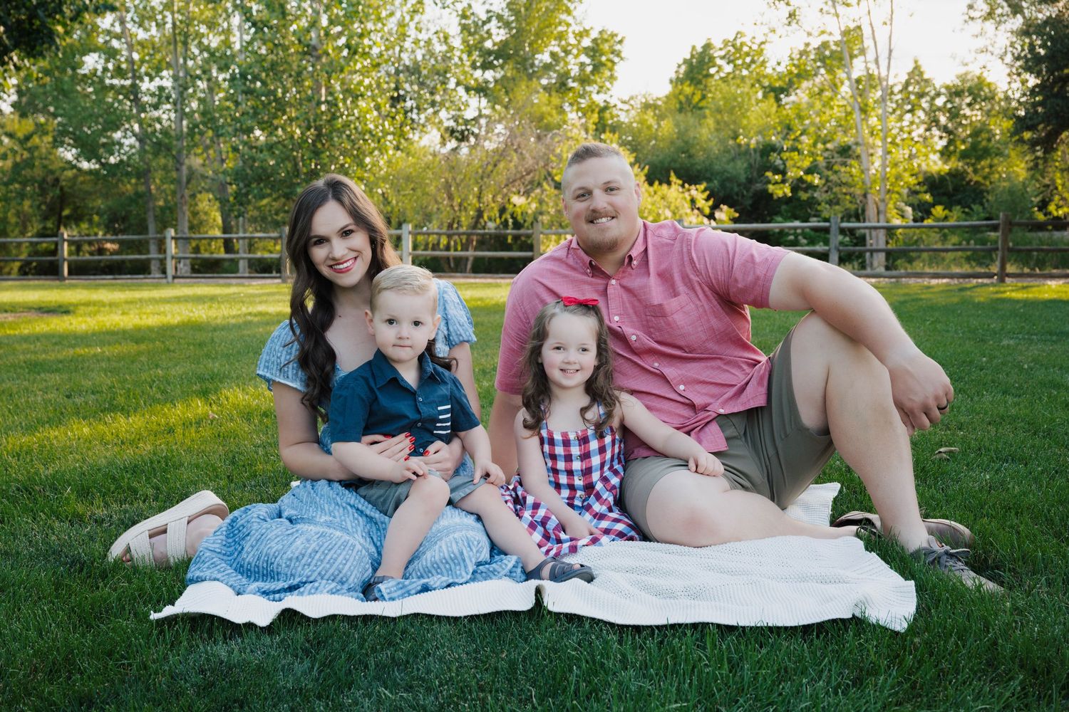 A family lounges together on a white blanket in a sunny park during a summer afternoon photo session.