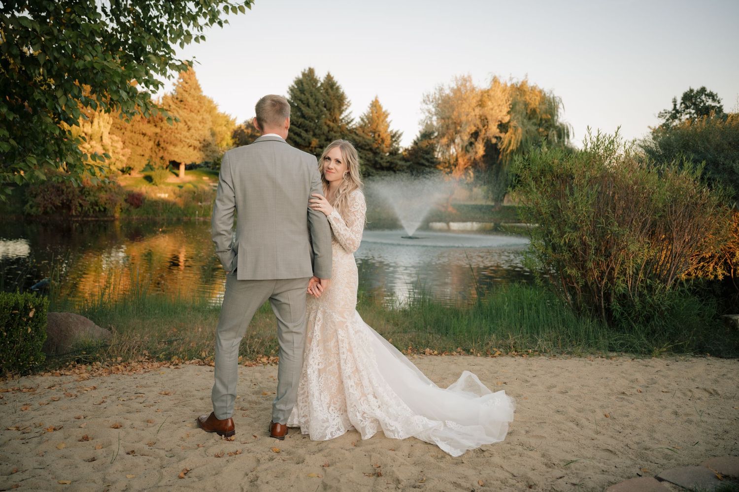 A couple stands together on a sandy beach near a pond with golden sunset light and fog creating a dreamy atmosphere.