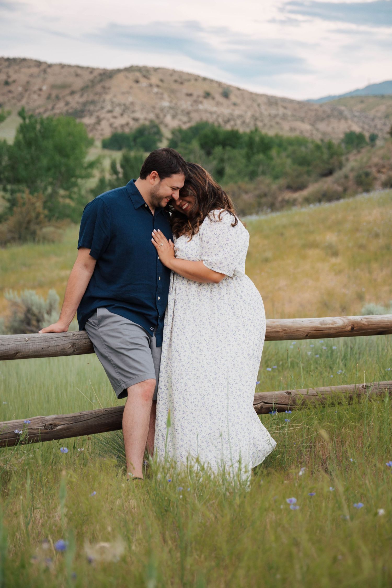 A couple shares an intimate moment together while sitting on a wooden fence in a scenic mountain meadow.