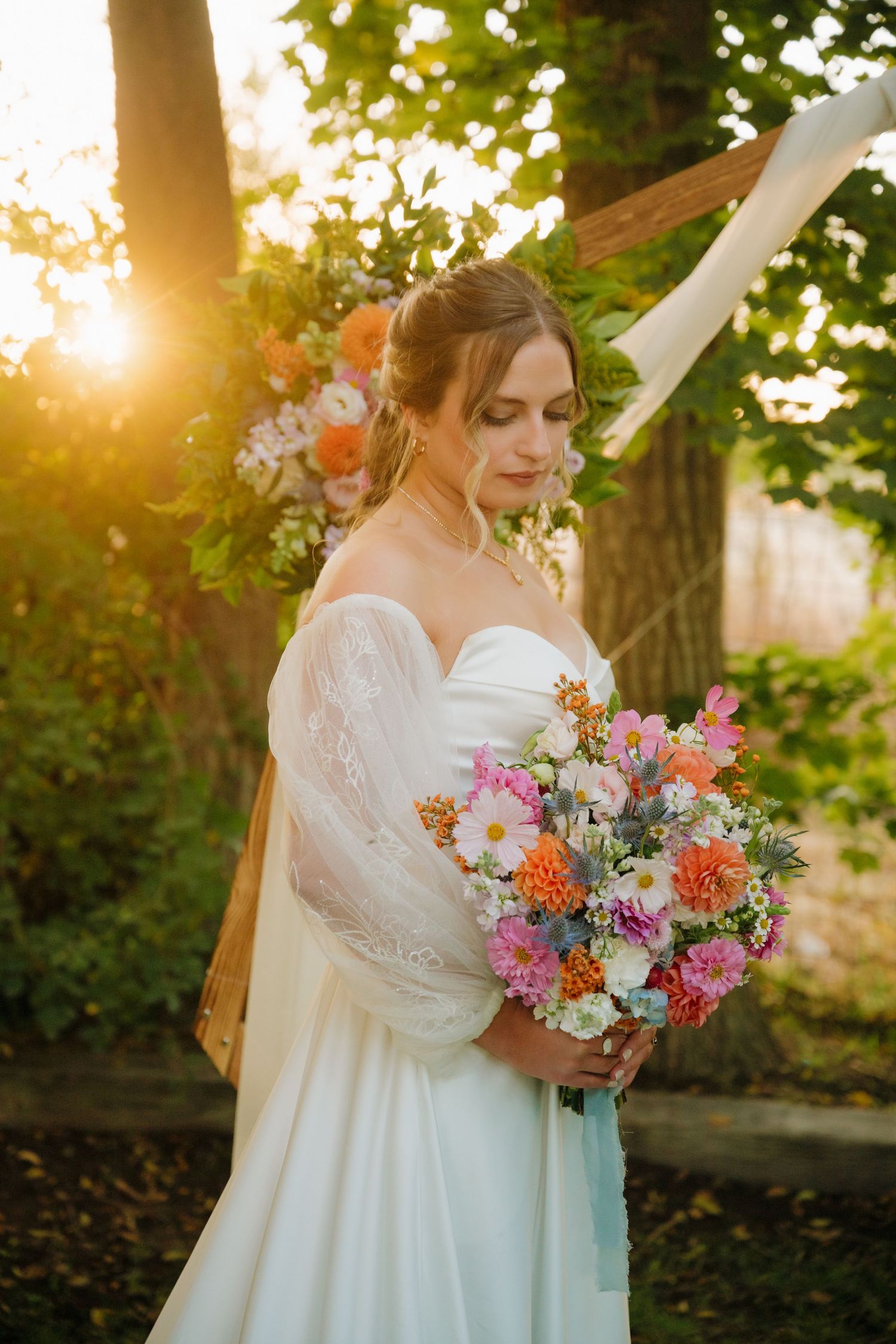 A bride holding a colorful bouquet poses in golden sunlight during sunset.