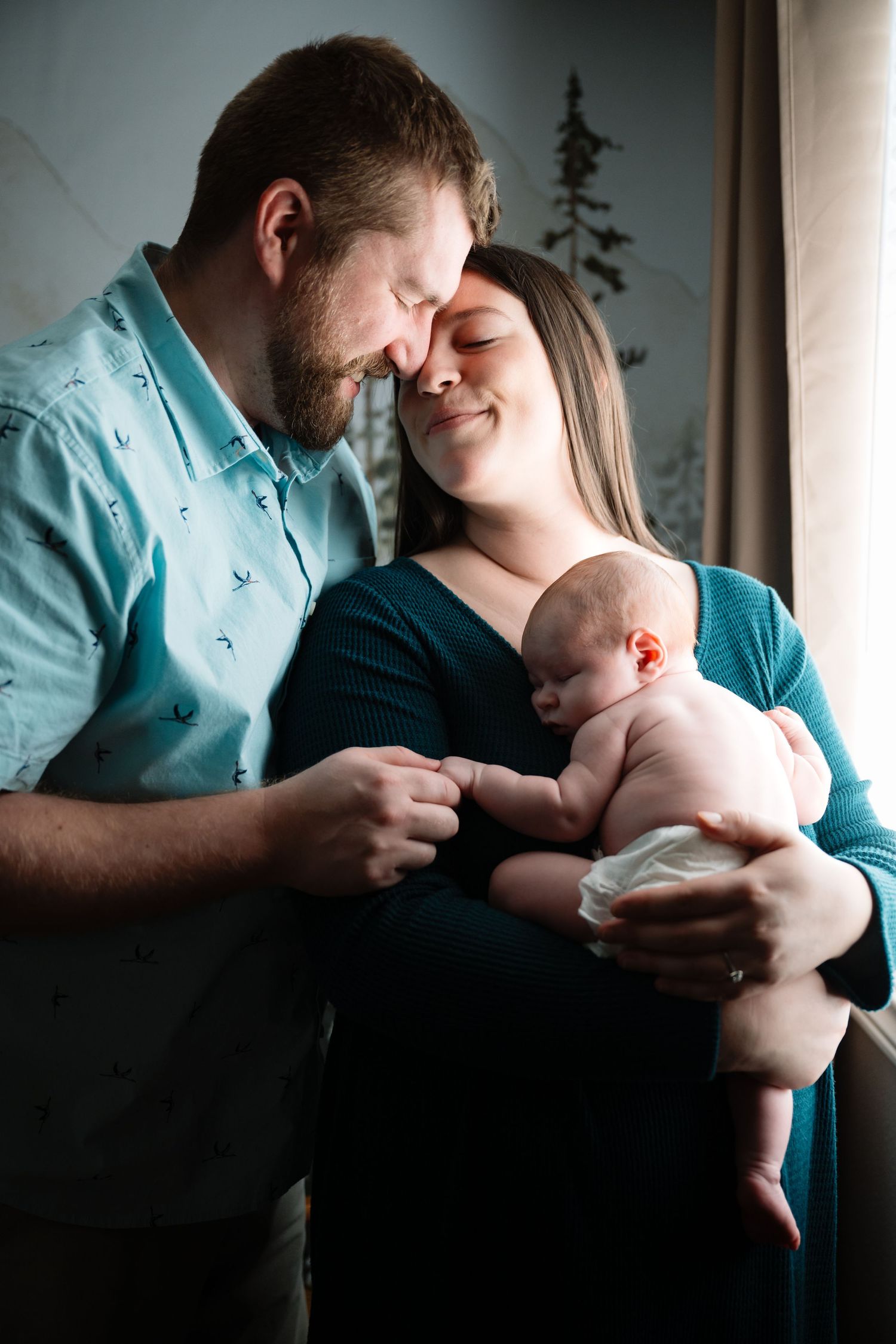 Tender family moment with newborn baby in nursery against window light.