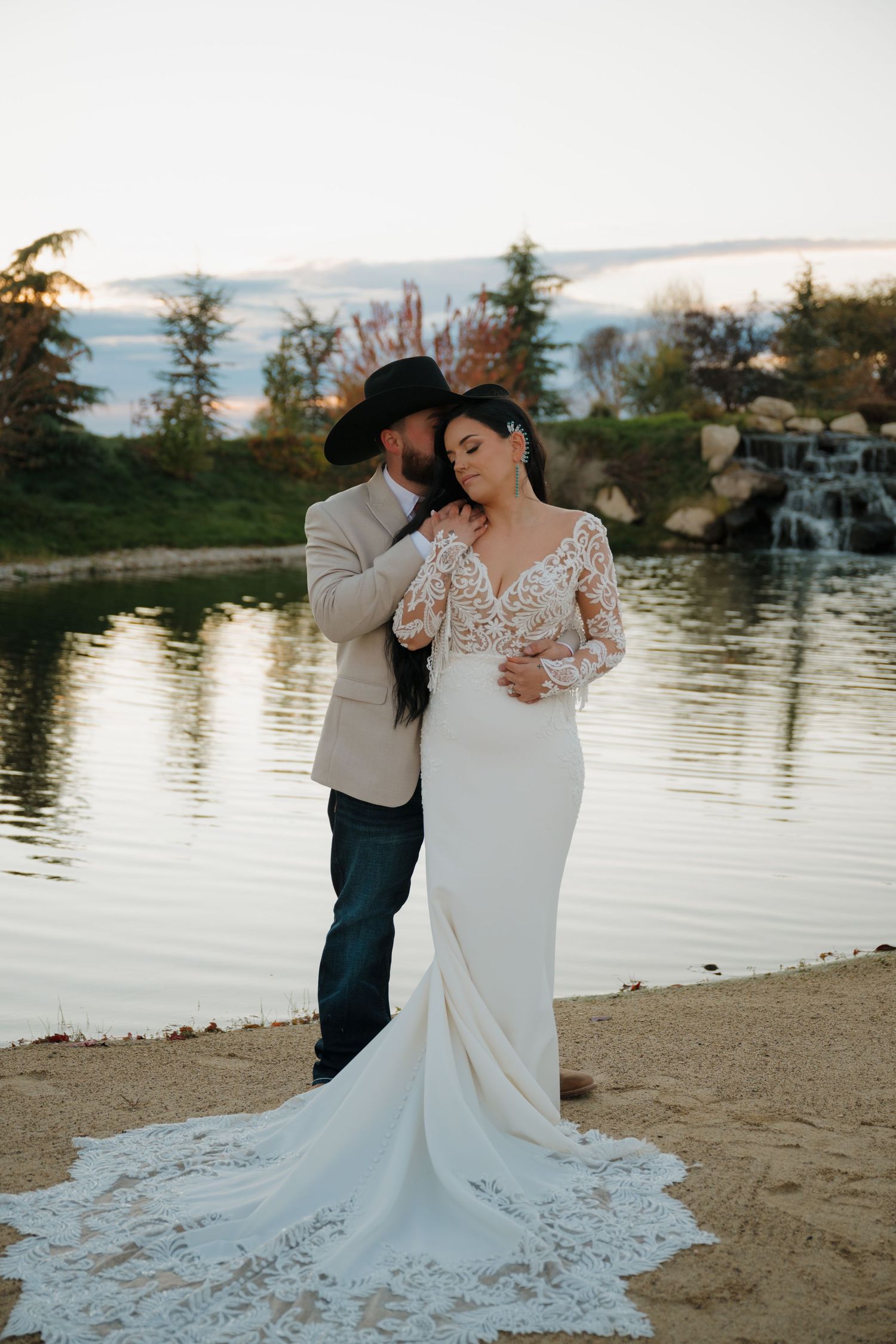 Romantic waterfront wedding photo with bride in lace dress and groom in cowboy hat.