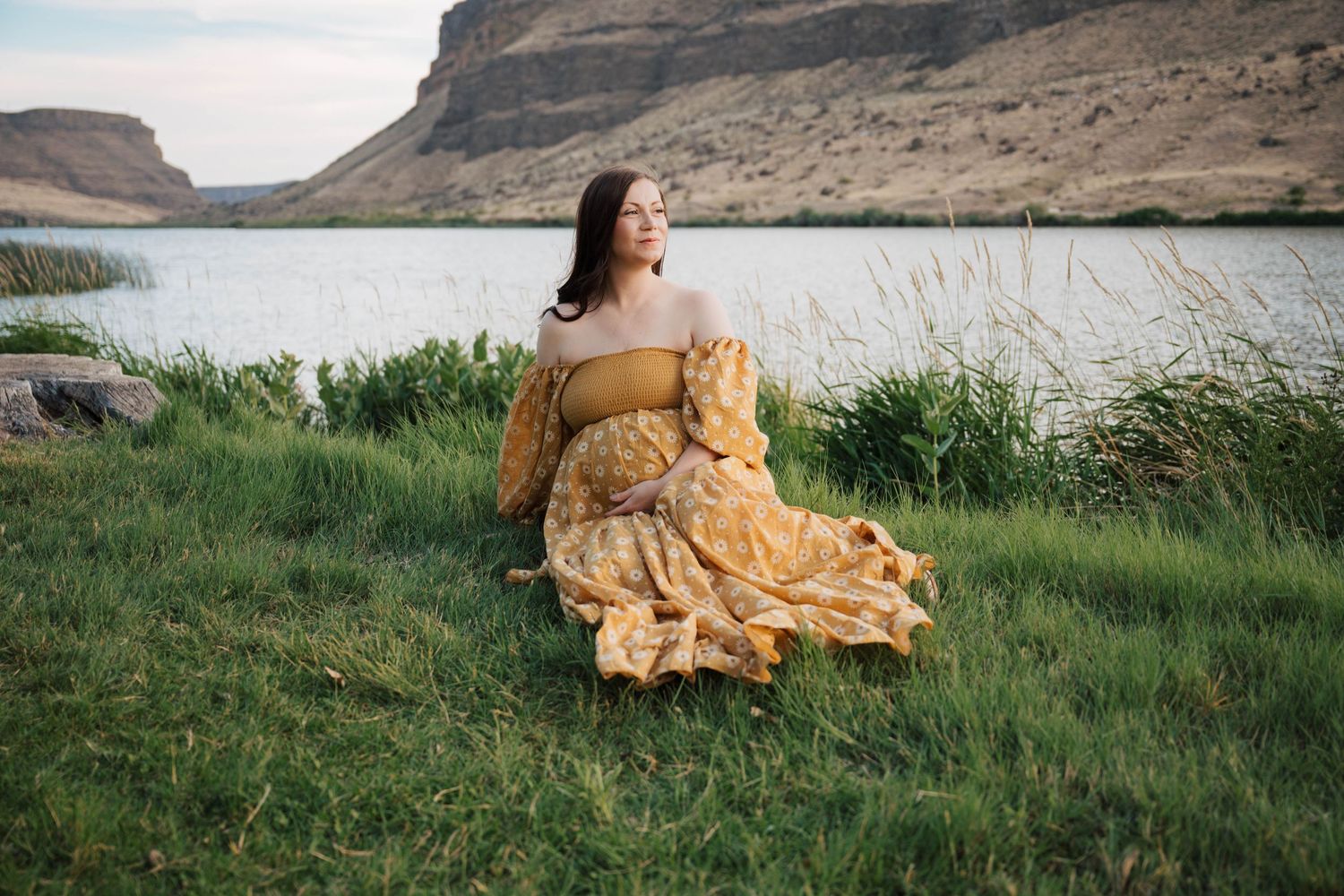 A person in a flowing yellow dress sits on grass by a scenic lake with rugged cliffs in the background.