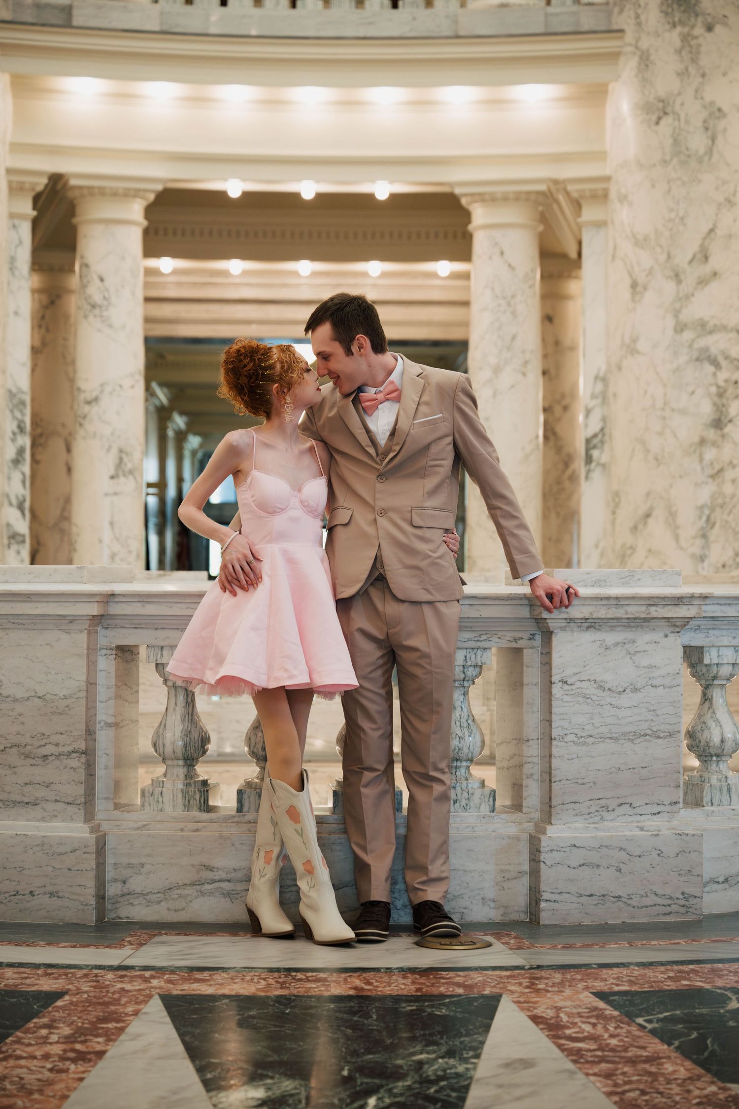 A romantic couple in cowboy boots and formal attire pose together in an elegant marble columned hallway.