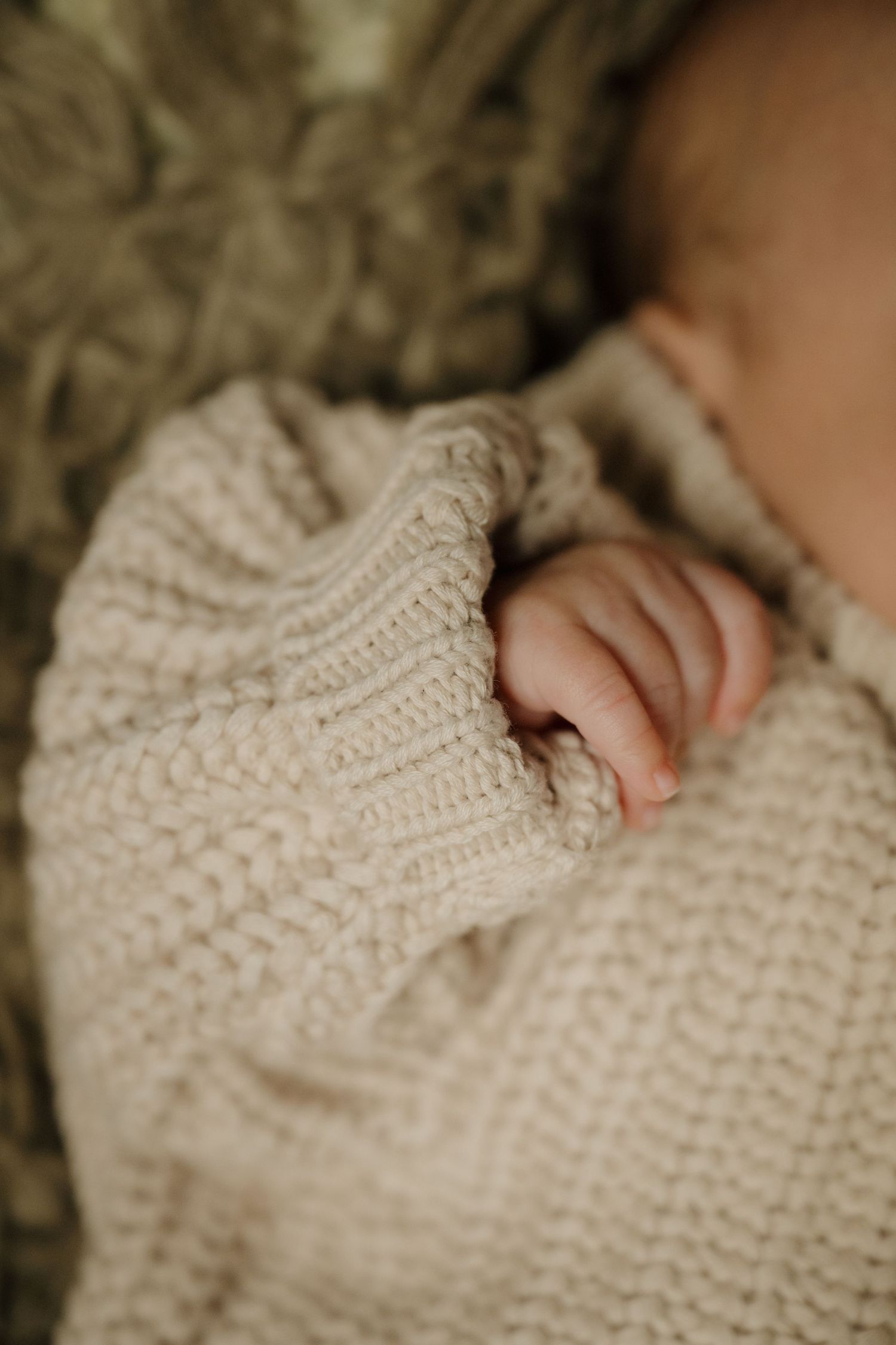 A tiny baby hand reaches out from under a cream knitted blanket.