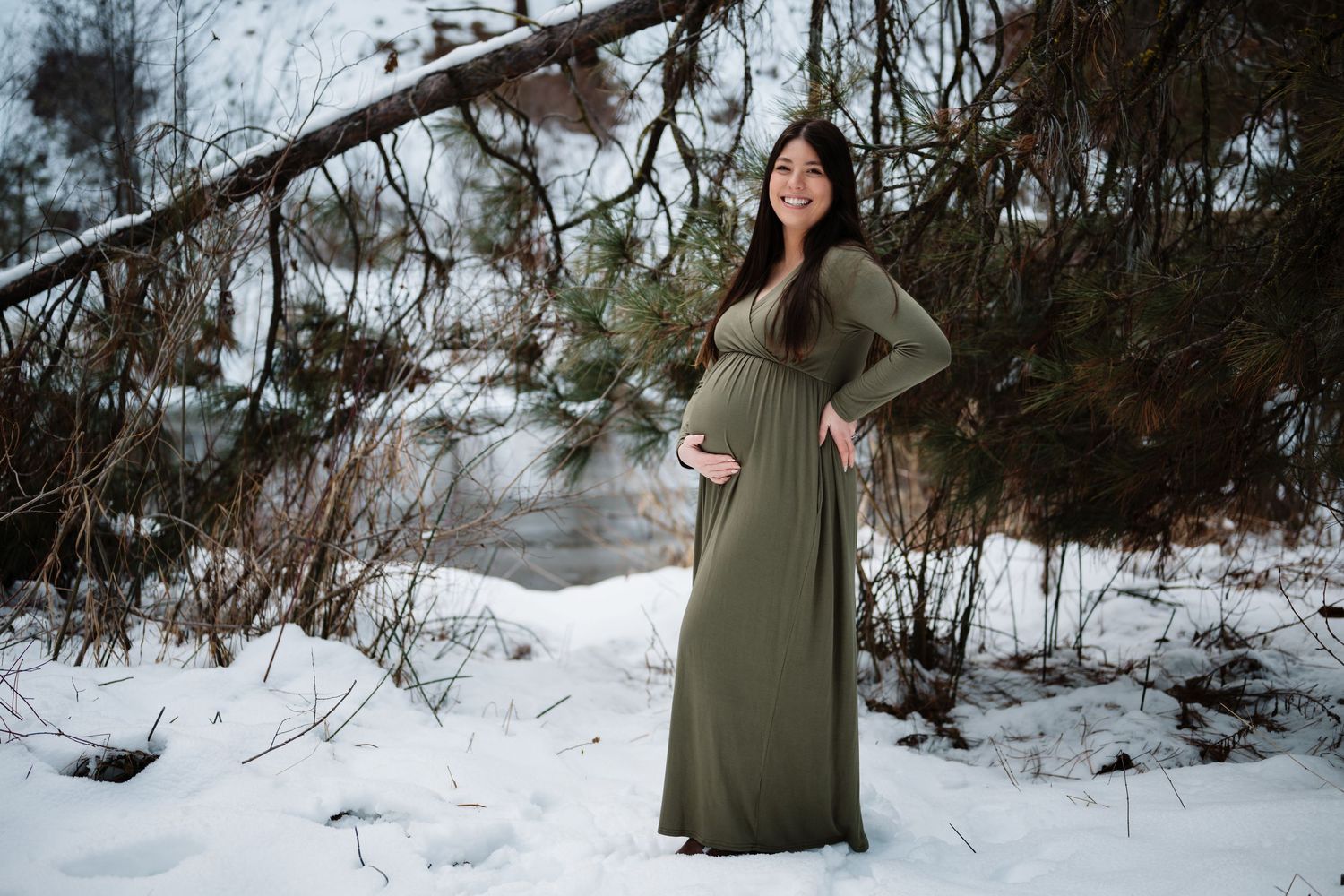 Winter maternity photo of woman in long olive dress against snowy forest landscape.