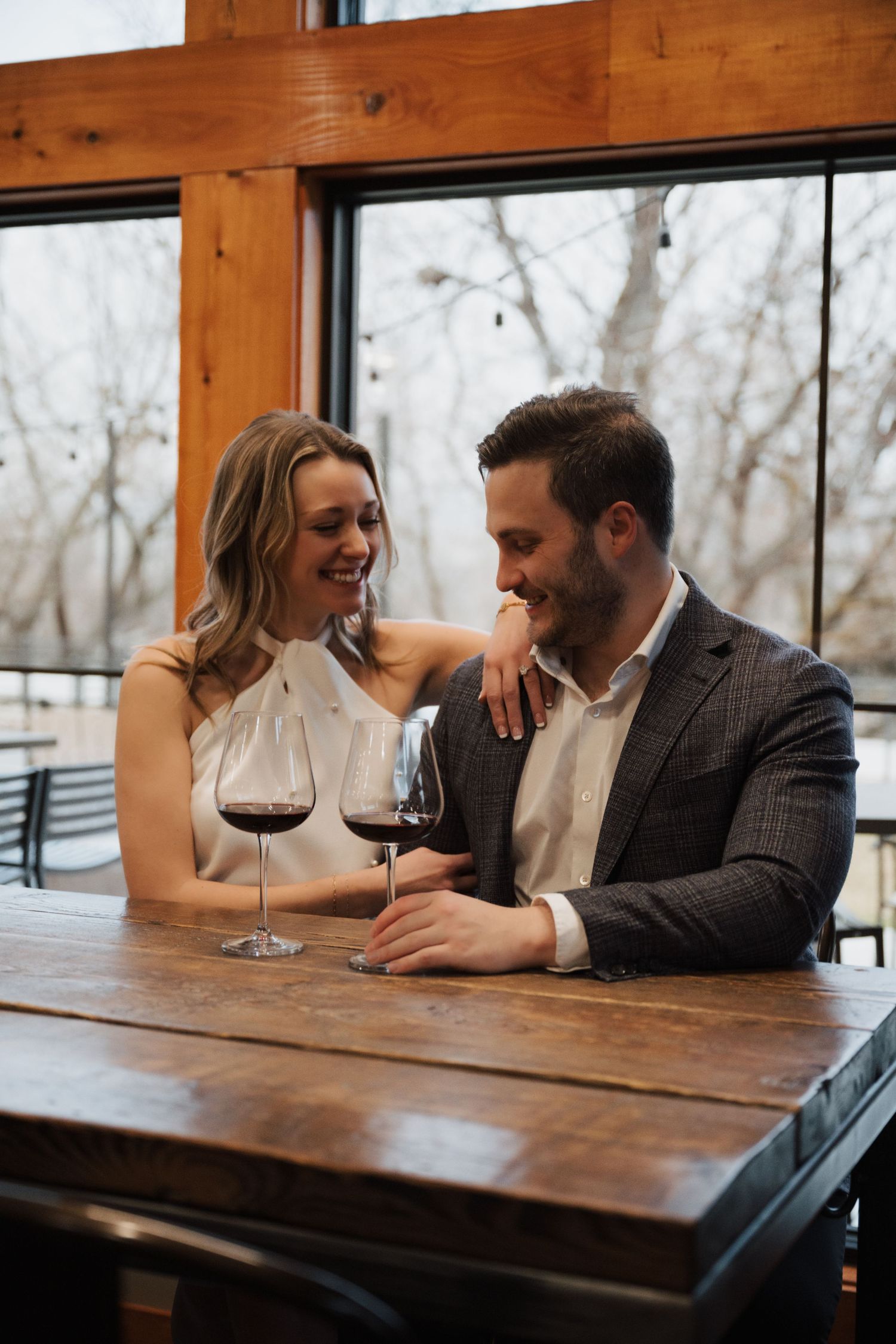 Couple sharing wine and laughter at rustic wooden table by large windows.