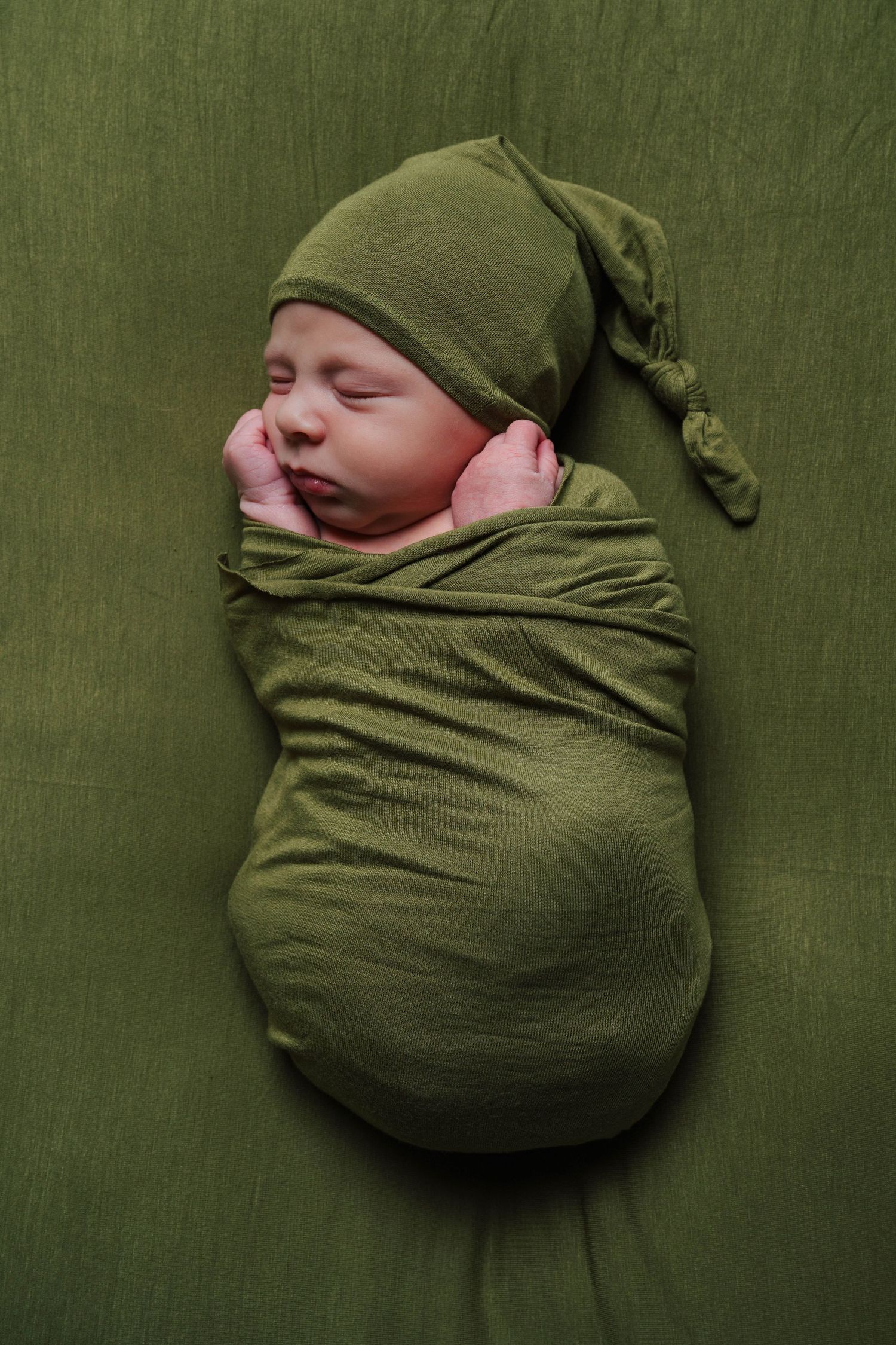 A sleeping newborn wrapped in olive green swaddle and matching hat on green textured background.