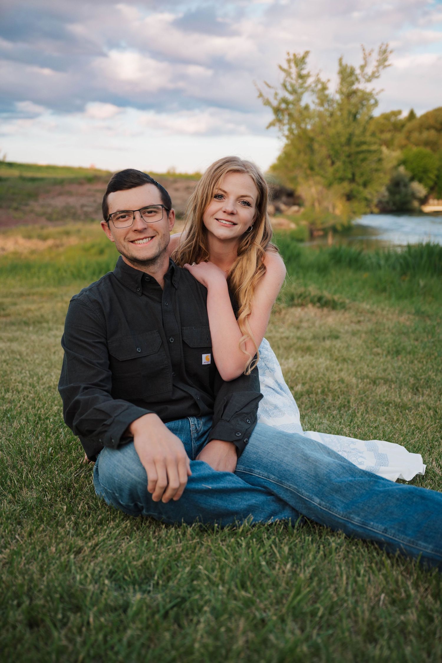A couple sits together on green grass during a golden hour outdoor photo session near a pond.