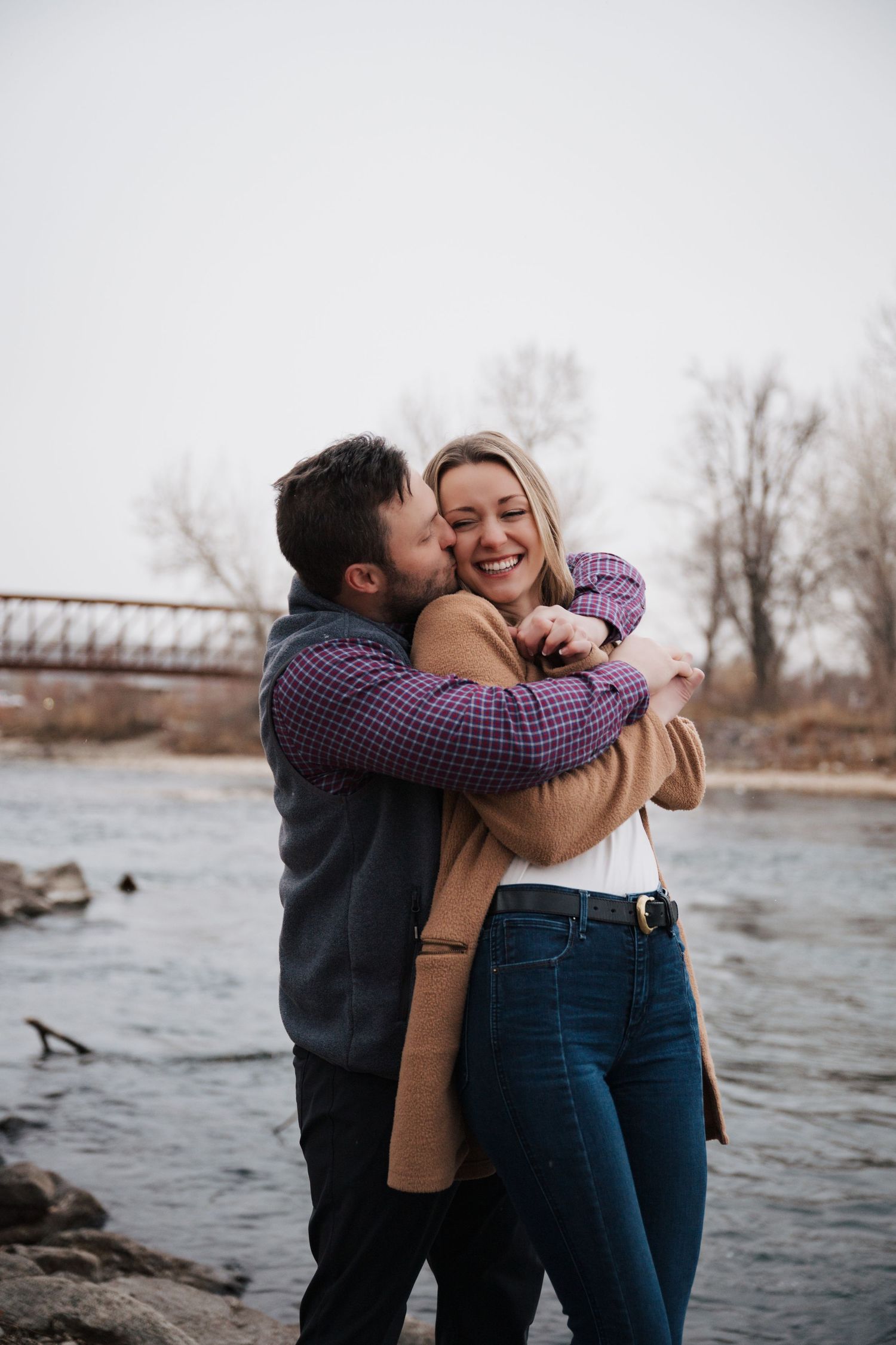 Candid embrace by riverbank with rustic bridge in background on cloudy day.