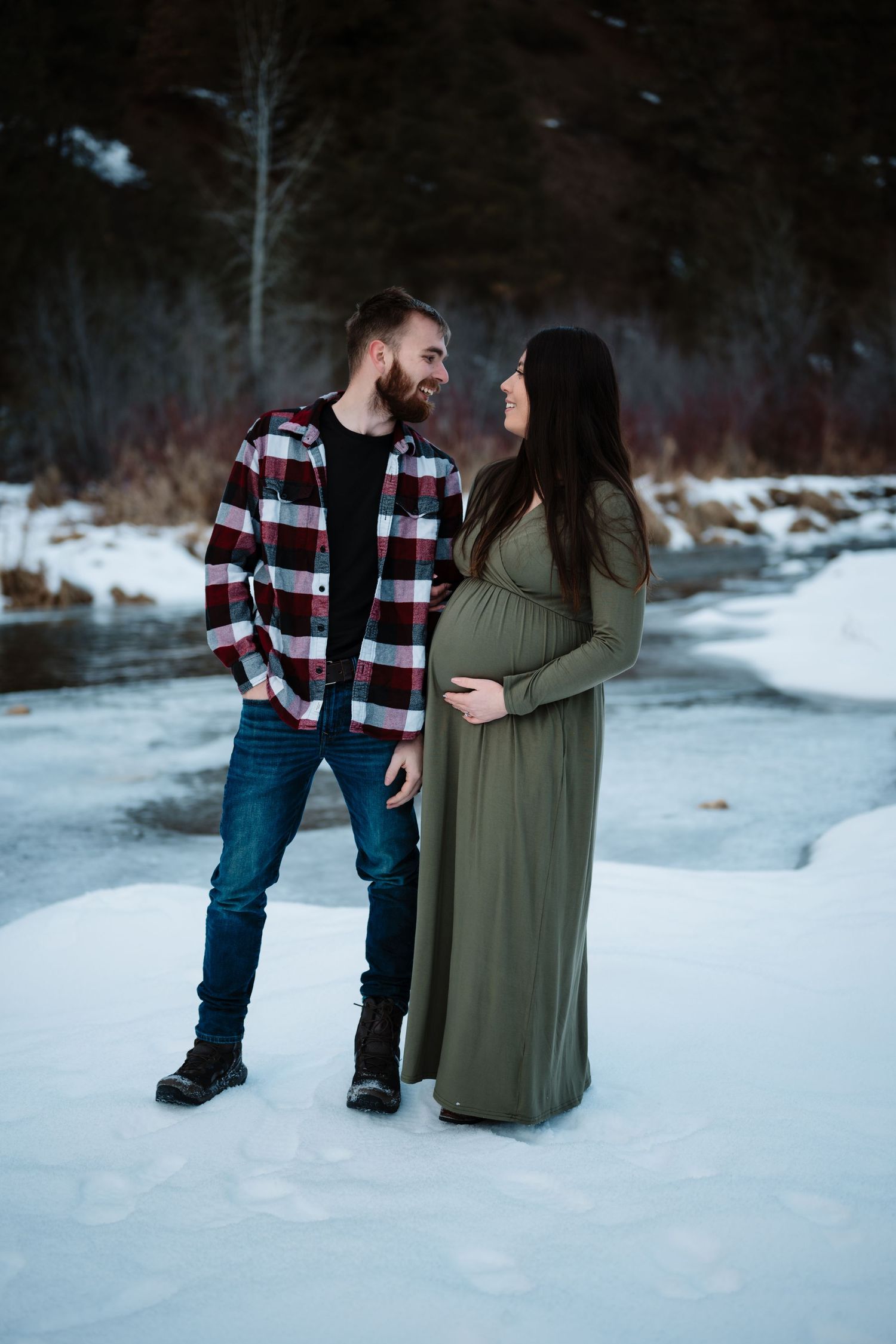 A couple shares a moment in the snow, with the woman wearing a flowing green maternity dress.