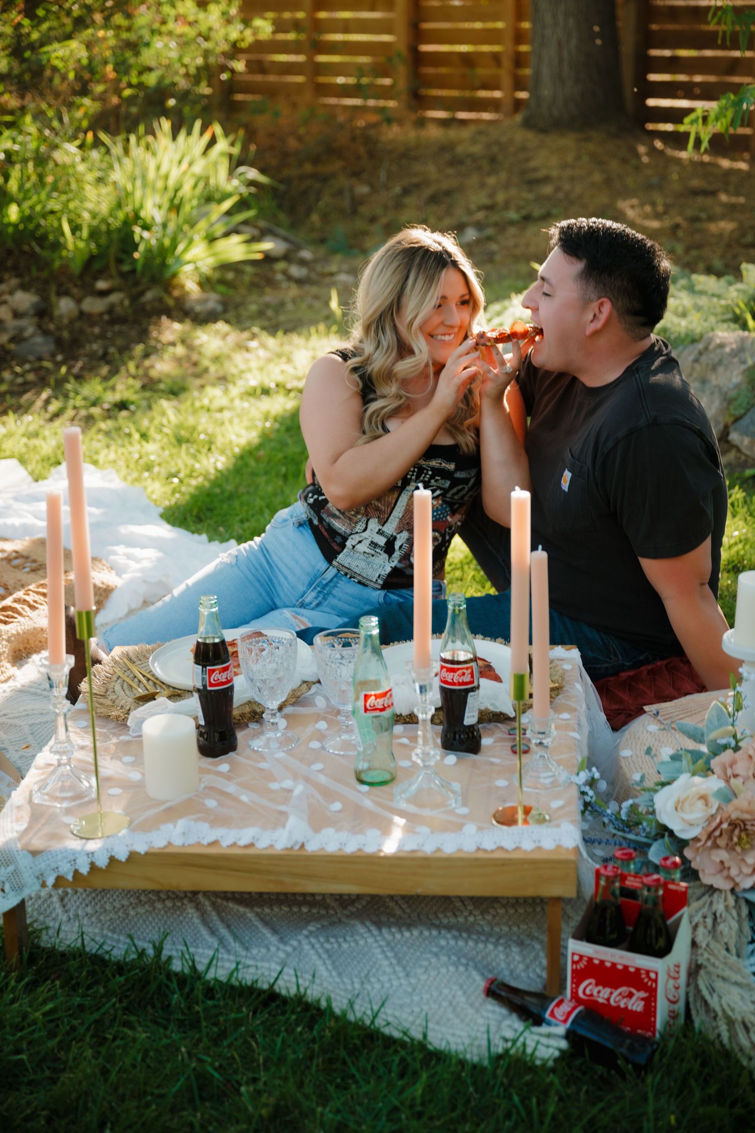 Couple playfully sharing food at a decorated outdoor picnic with Coca-Cola and candles.