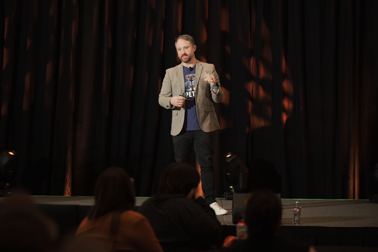 A performer in a gray blazer stands on stage delivering comedy in front of a dark curtain backdrop at a nightclub.