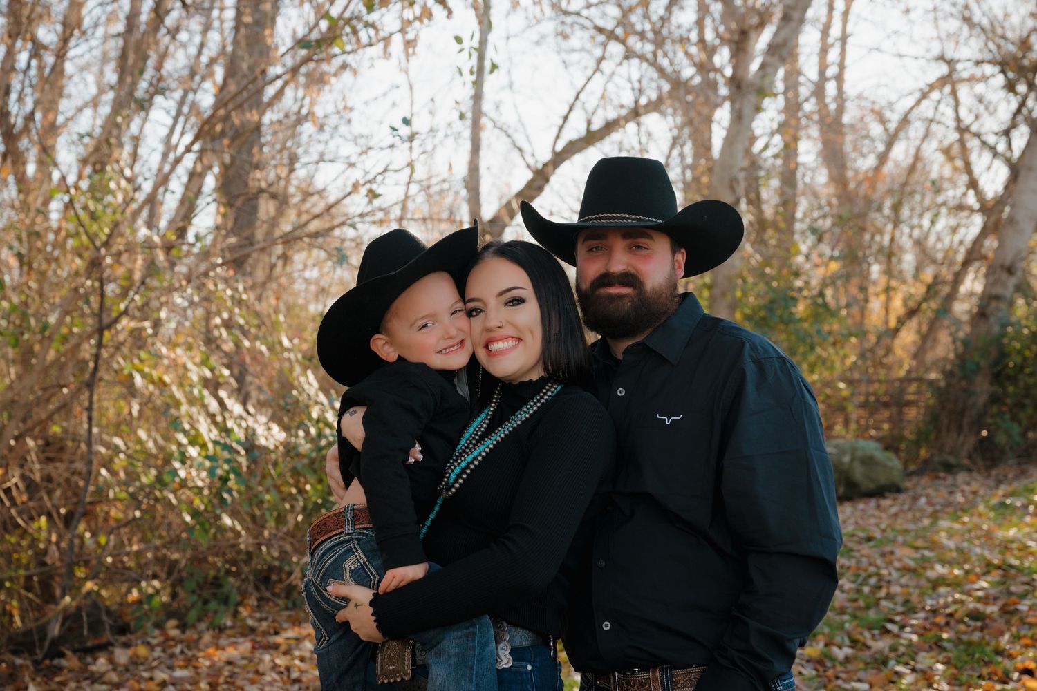 A family in western wear poses together outdoors during autumn among fallen leaves and bare trees.