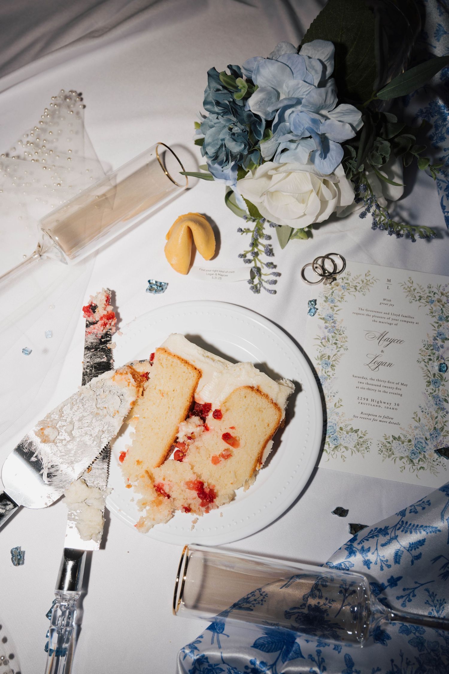 A wedding table setting with sliced cake, blue and white flowers, rings, and invitation card on a white surface.