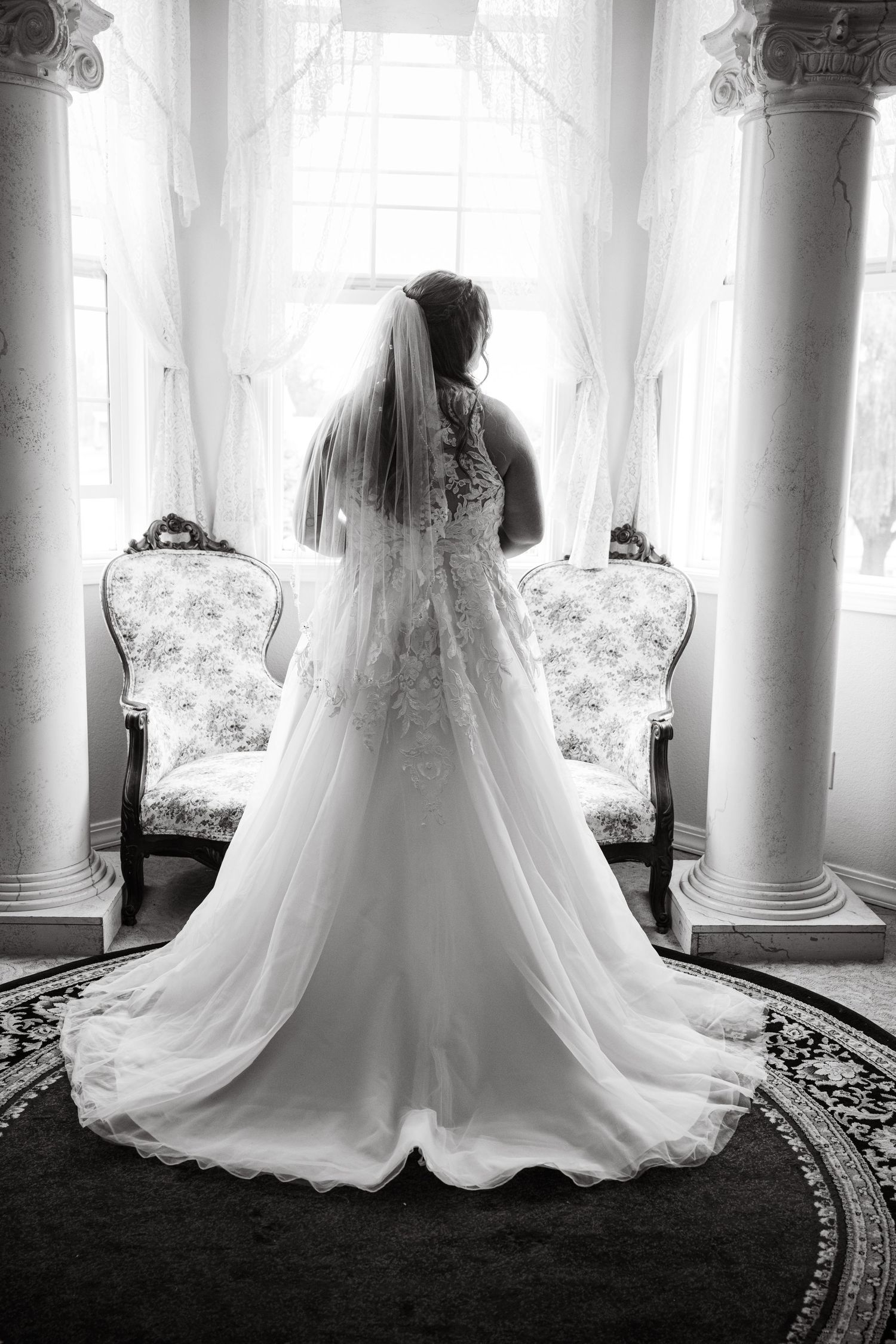 Back view of a bride in an elegant lace wedding gown standing between ornate columns near a window.