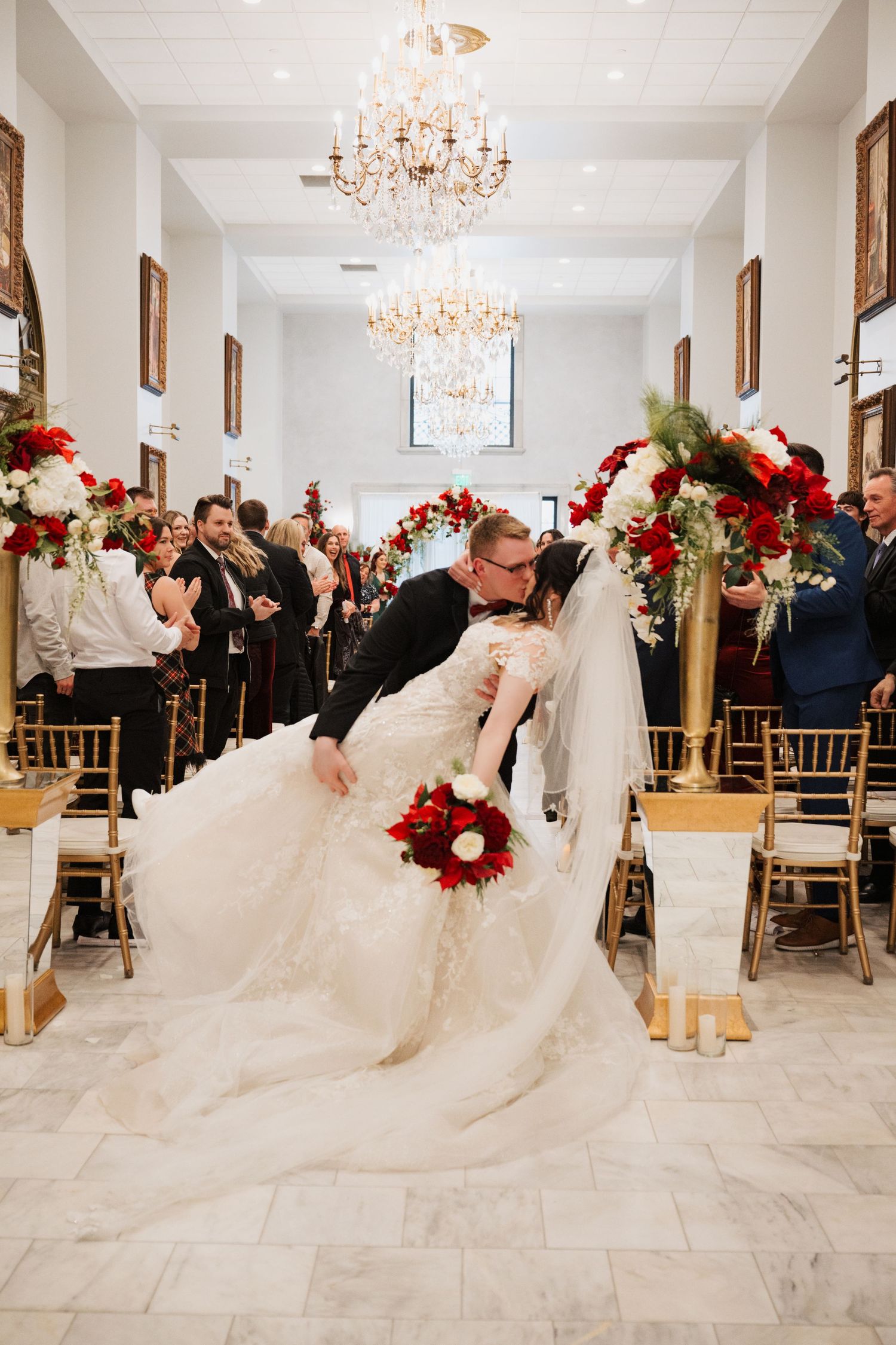 Dramatic wedding dip kiss under crystal chandeliers with red floral decorations.