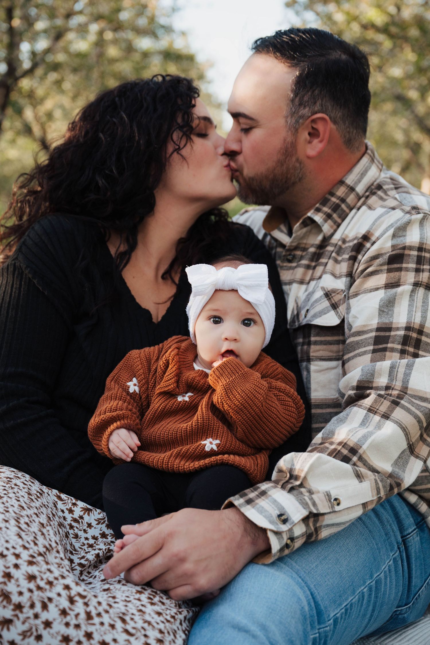 Couple kissing outdoors with baby in rustic brown sweater and white bow headband between them during autumn photoshoot.