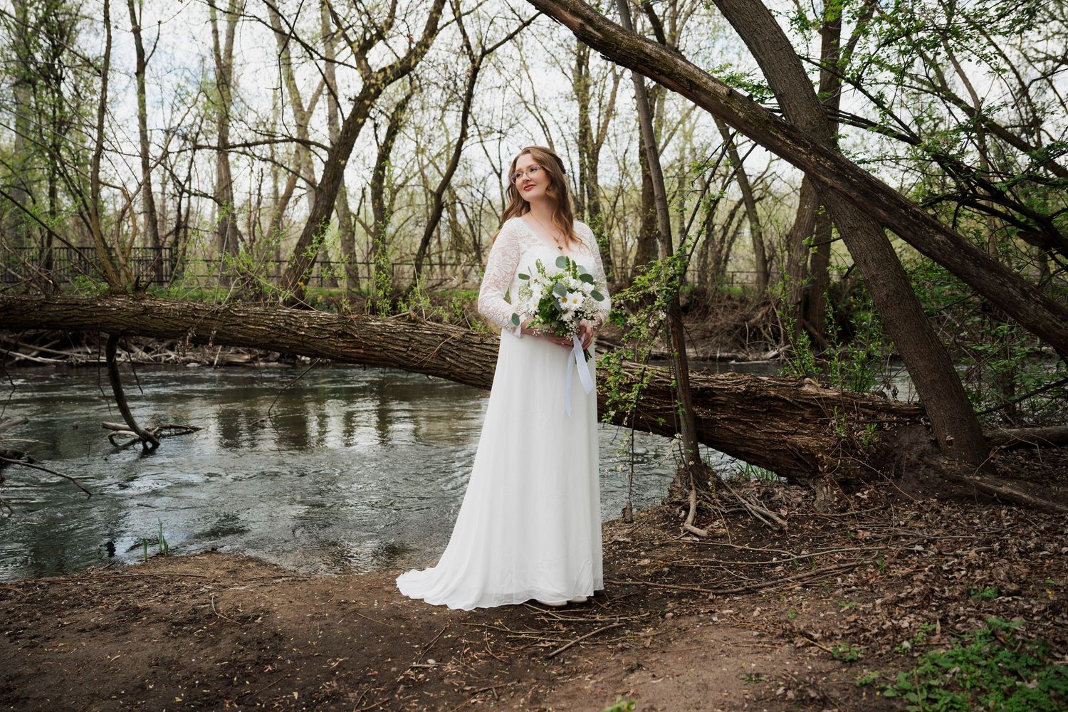 A bride in a white lace dress poses near a flowing stream in a wooded forest setting.