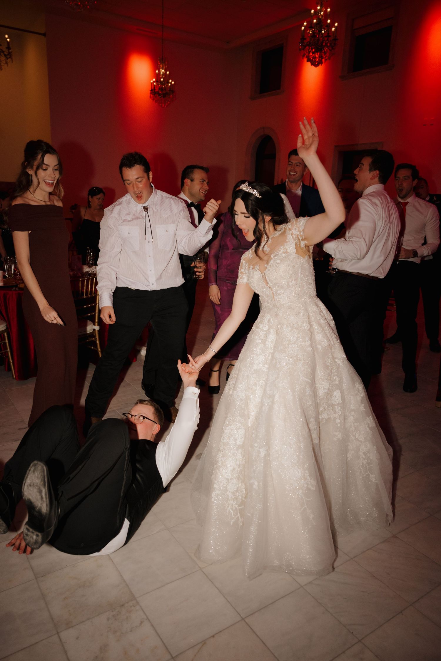 Wedding reception dance party with guests dancing energetically on a tiled floor under dramatic red lighting.