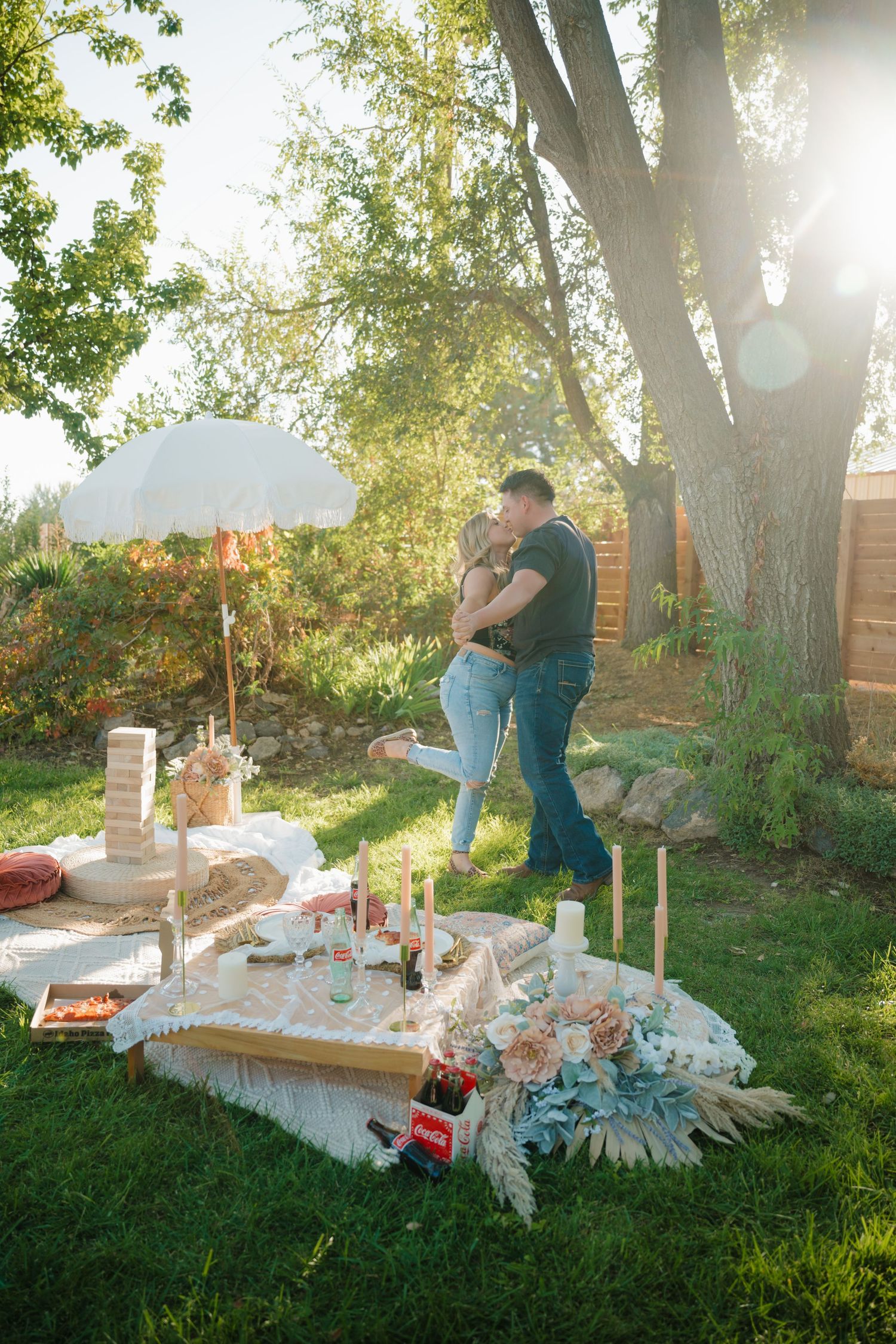 A romantic picnic setup in a backyard with candles, flowers and Coca-Cola bottles under trees.