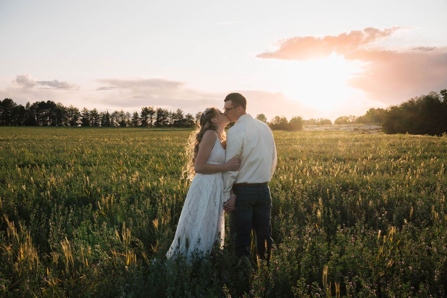 Silhouetted couple stands in golden wildflower field at sunset with orange sky and tree line in background.