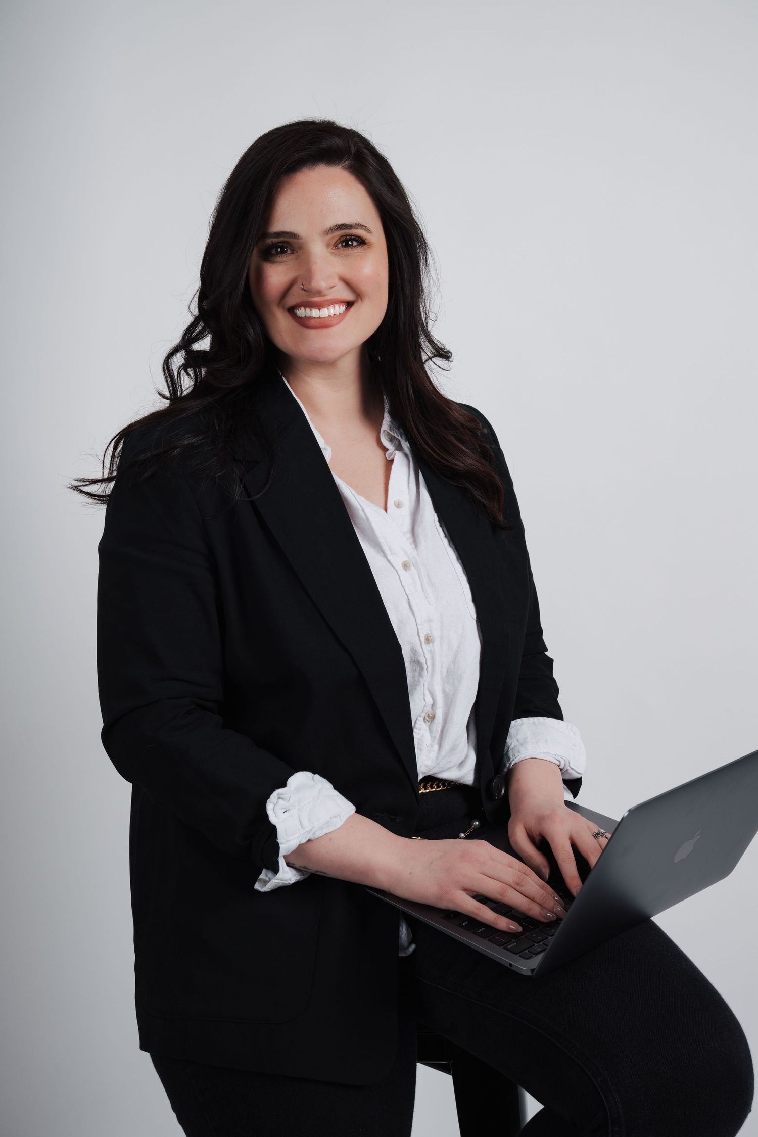 Professional business portrait of a person in black blazer and white blouse holding laptop against light gray background.