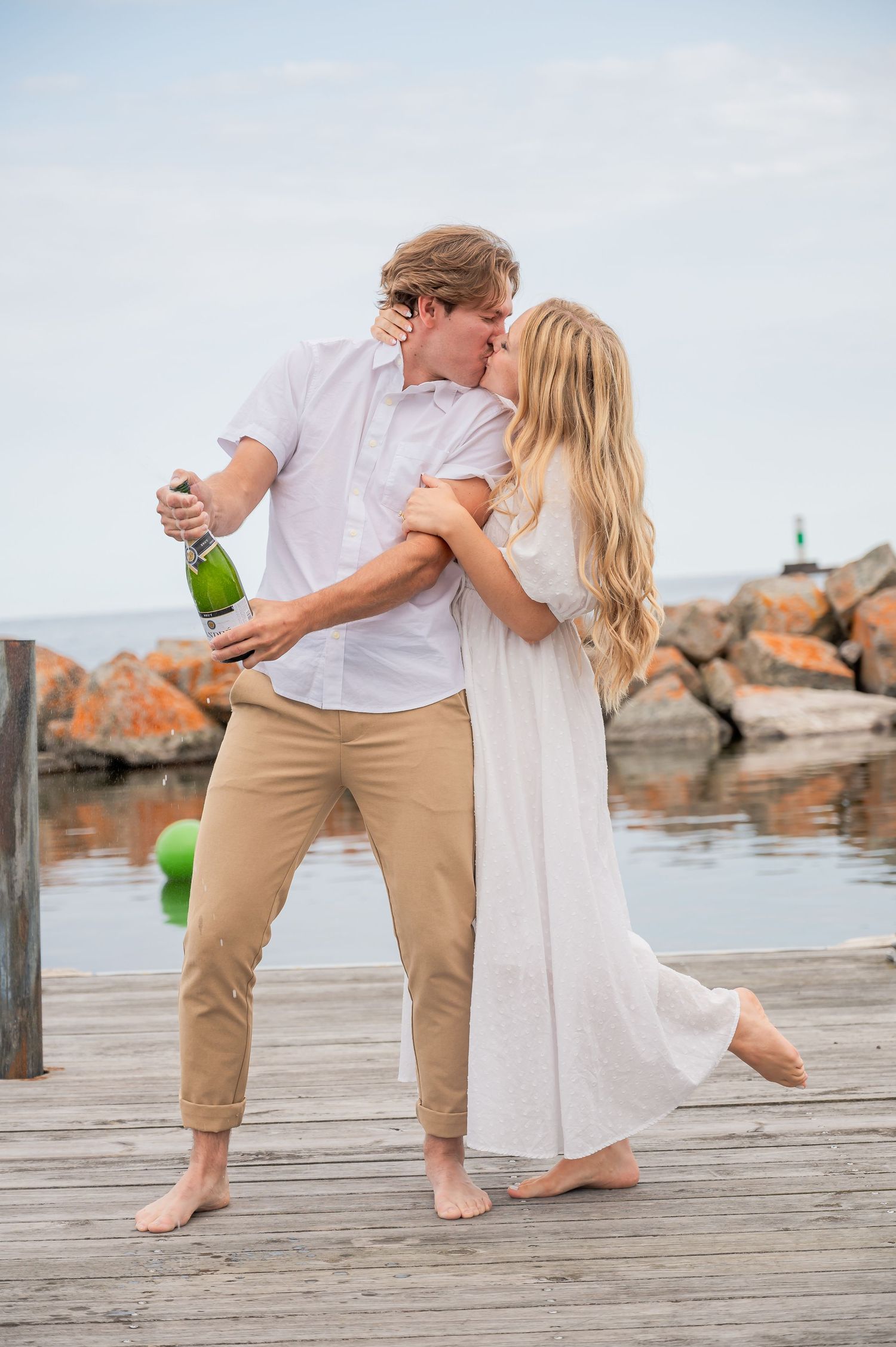 A couple celebrates with champagne on a wooden dock by the water, embracing barefoot in casual summer attire.