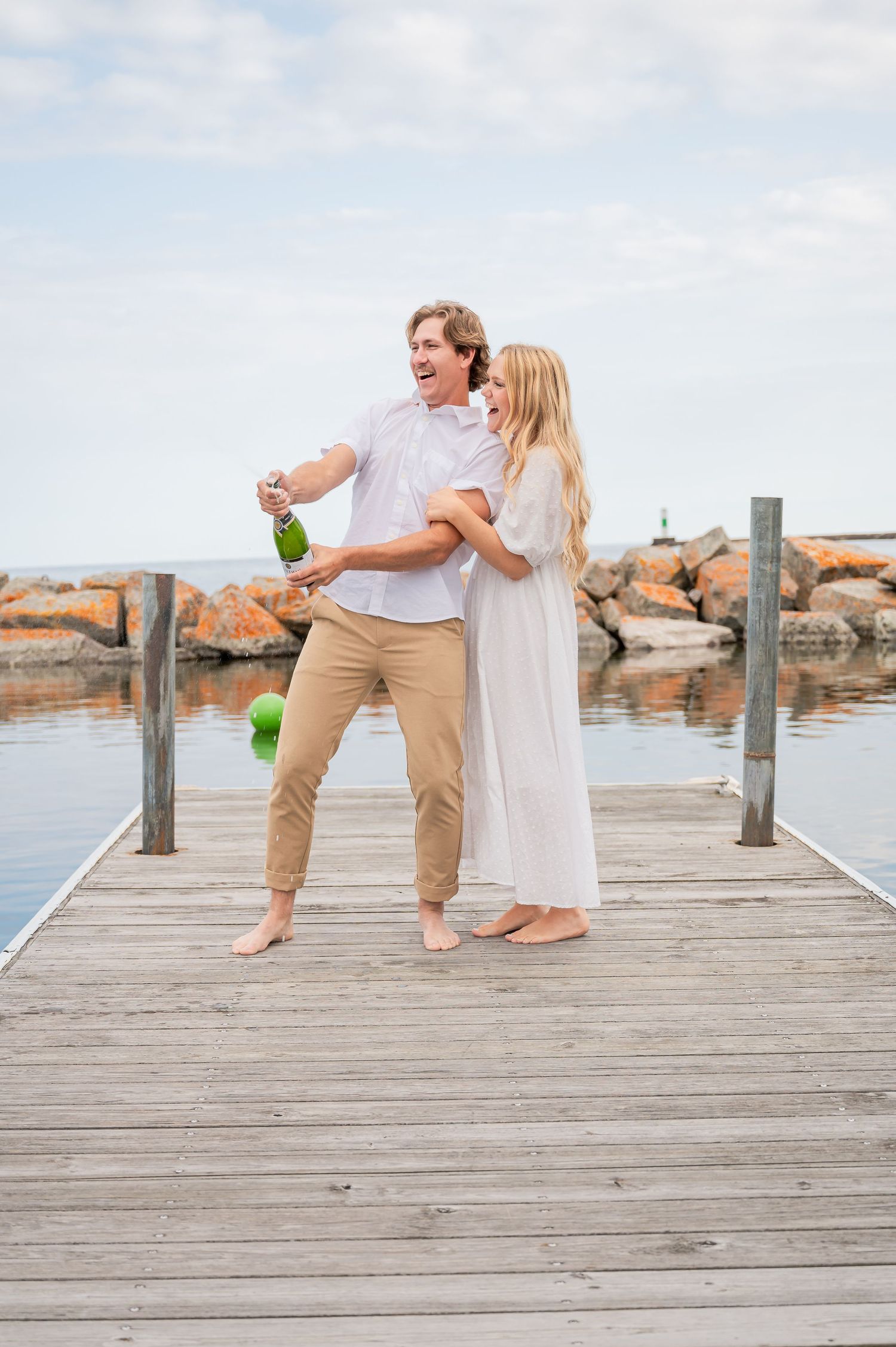 A joyful moment between a couple popping champagne on a wooden pier by the ocean on a sunny day.