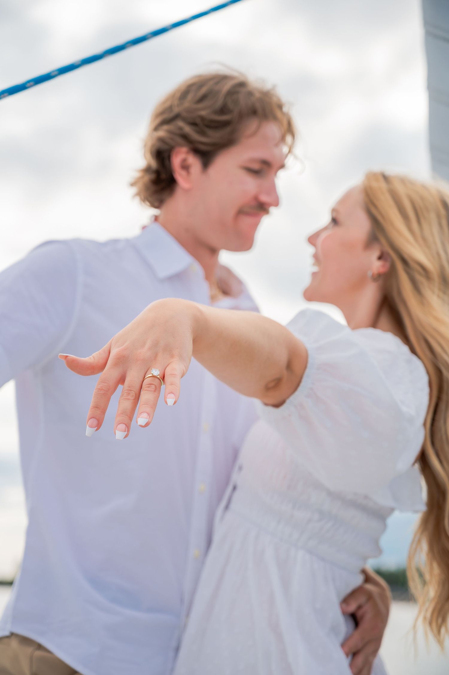 A couple in white clothing poses together on a boat while showing off a new engagement ring on display.