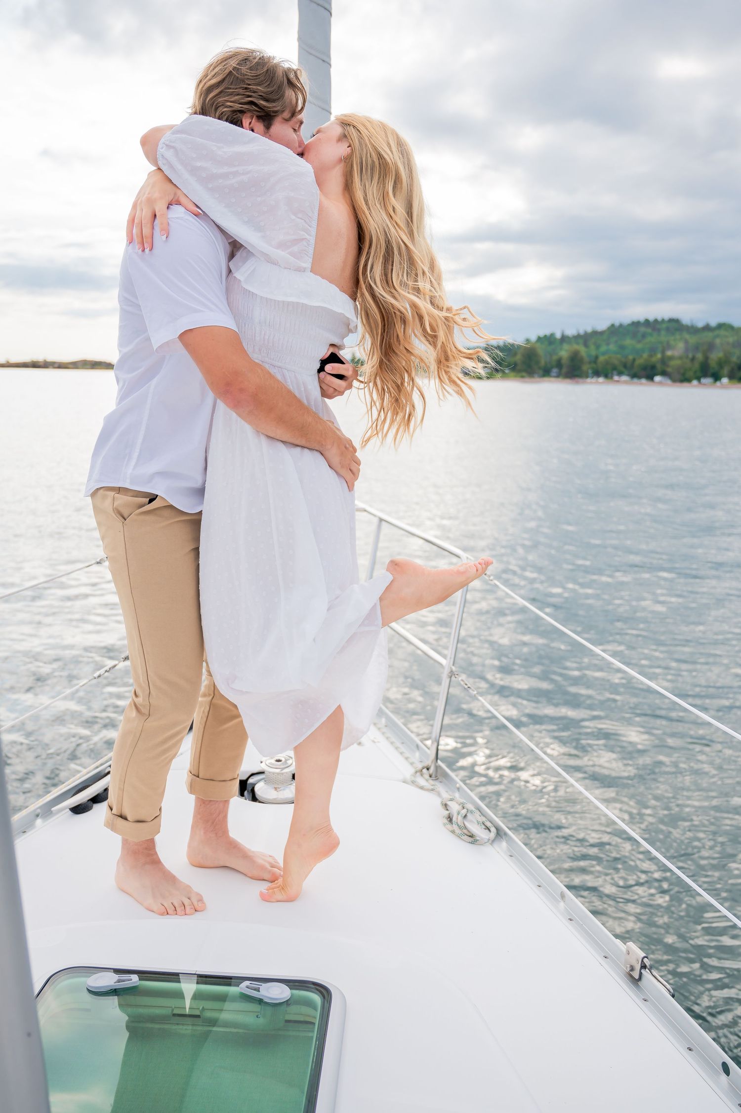Romantic couple embracing and kissing on the bow of a sailboat on a calm lake with cloudy sky background.