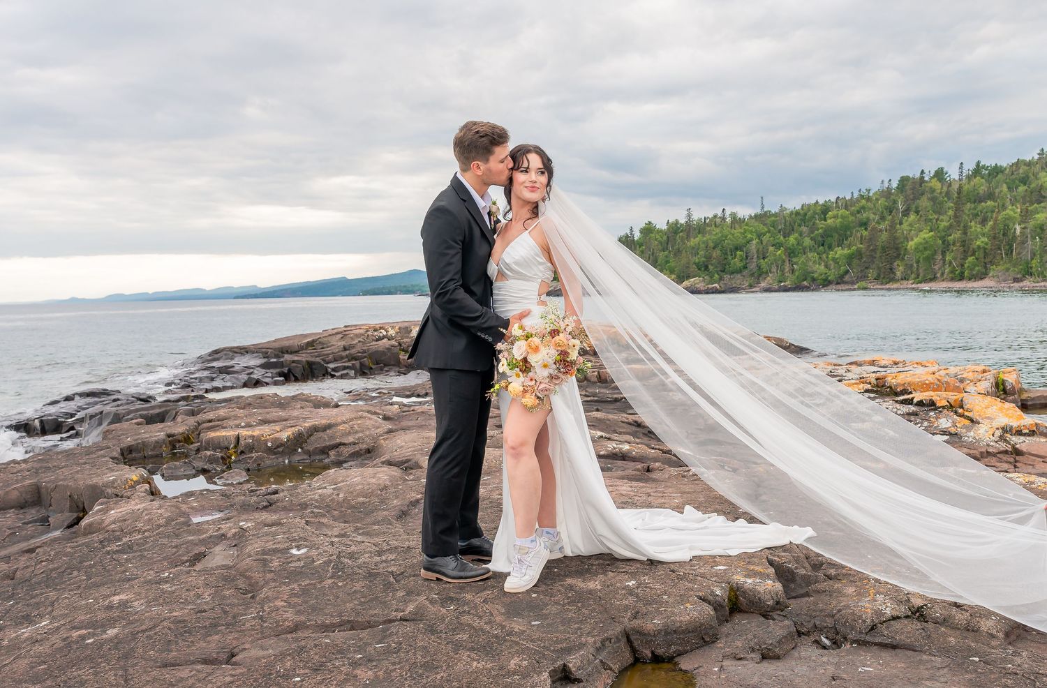 A scenic lakeside wedding portrait with a dramatic veil flowing in the wind on rugged coastal rocks.